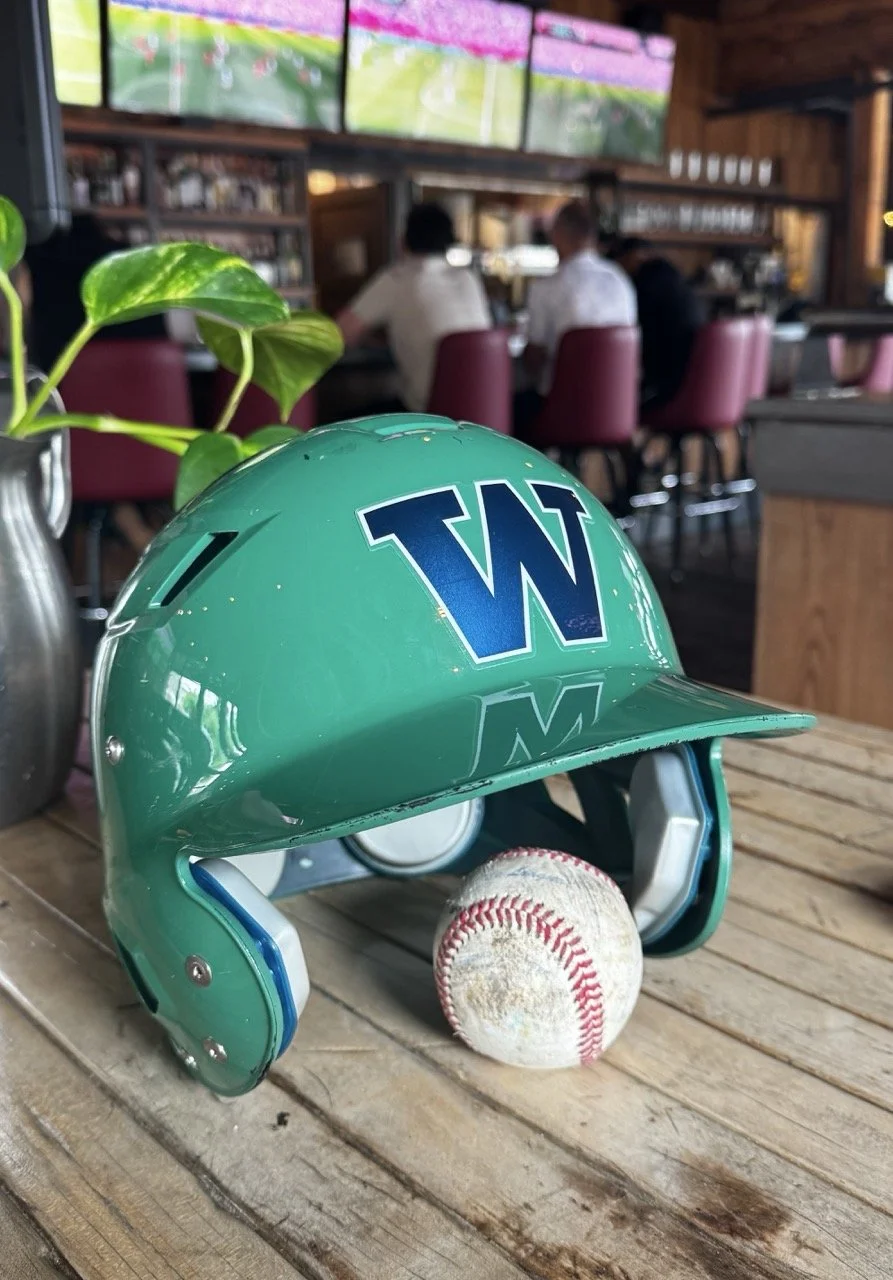A teal baseball helmet with a blue 'W' logo on it, placed on a wooden table with a baseball underneath it, in a sports bar setting with televisions and people in the background.