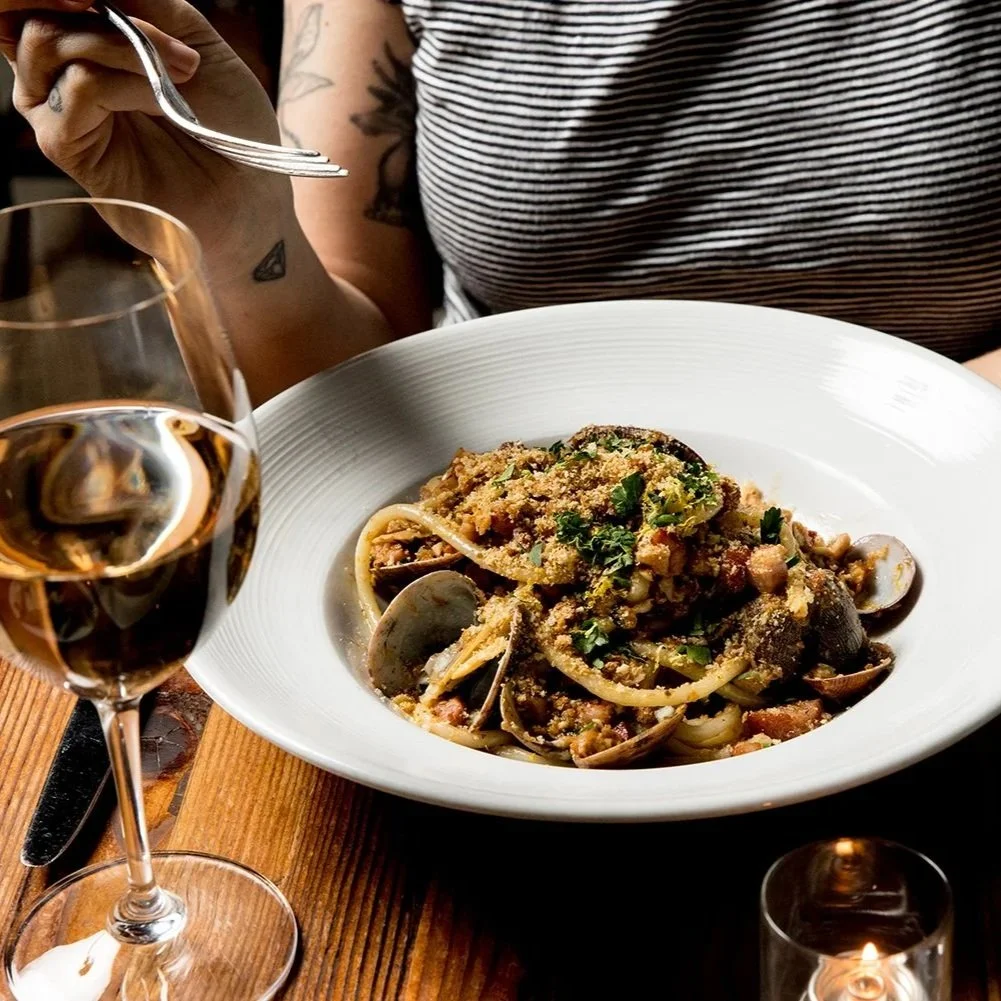A person in a striped shirt sitting at a wooden table with a large white bowl of pasta topped with herbs and breadcrumbs, along with two glasses of what appears to be rosé wine.