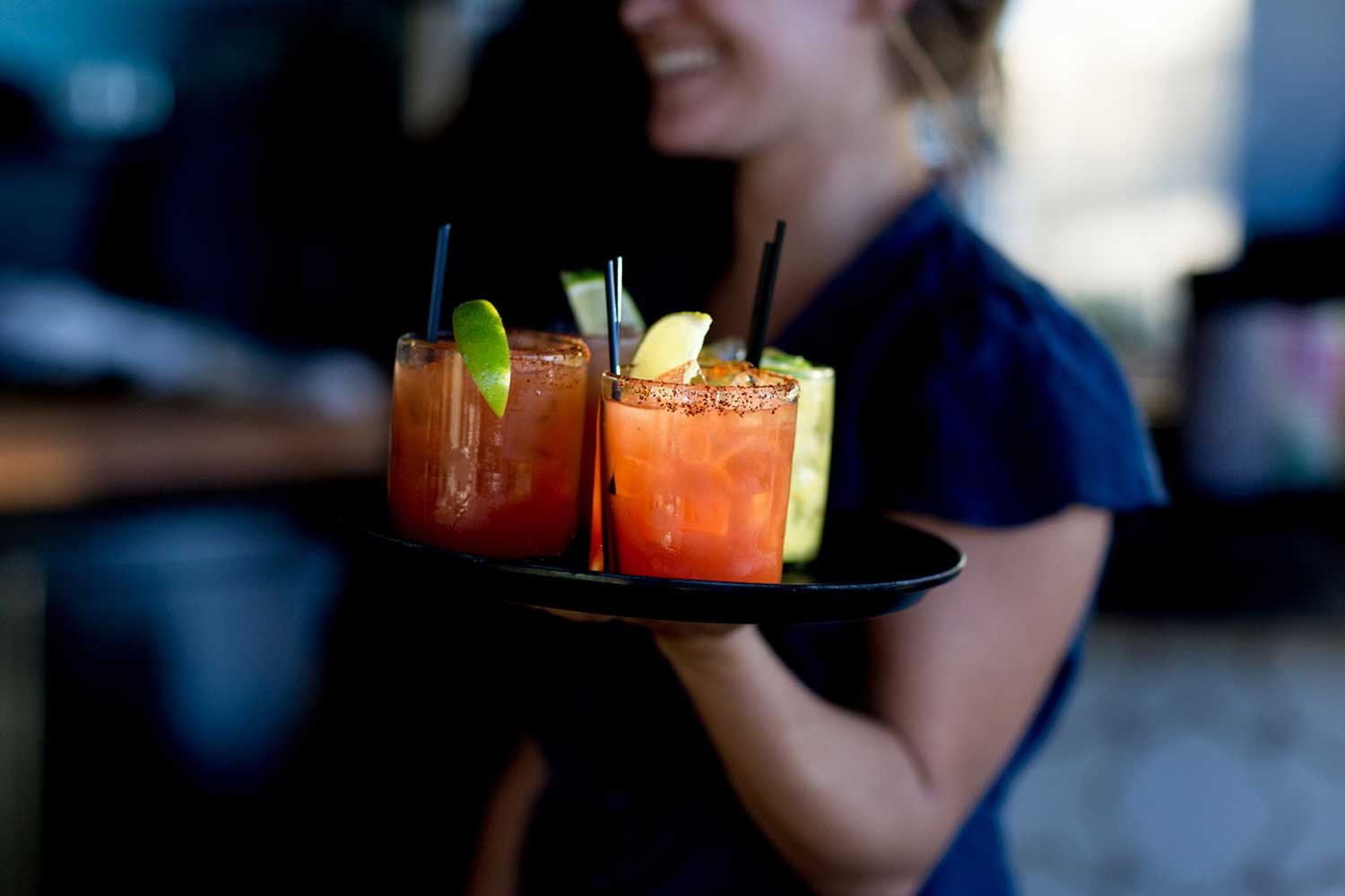 A woman holding a tray with three colorful cocktails garnished with lime and lemon slices, salt-rimmed glasses, in a dimly lit setting.