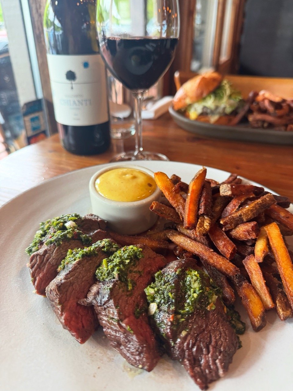 A plate of sliced beef tenderloin topped with chimichurri sauce, served with crispy French fries, a side of hollandaise sauce, a glass of red wine, and a bottle of red wine on the table in a restaurant.