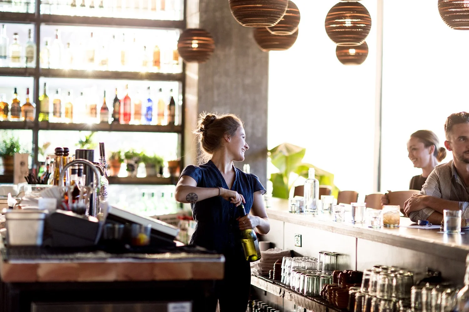 A smiling female bartender with a tattoo on her left forearm, holding a green beer bottle, standing behind a bar counter in a well-lit, modern restaurant or cafe with hanging globe pendant lights and shelves of liquor bottles in the background. Two p