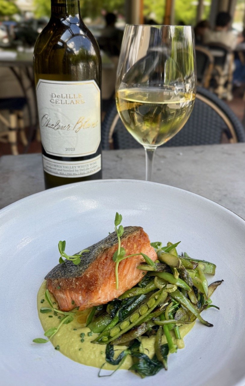 A plate of cooked salmon with green beans and sauce, garnished with microgreens. Behind it is a glass of white wine and a bottle of Chardonnay from DeLille Cellars. The setting is an outdoor restaurant with blurred people in the background.