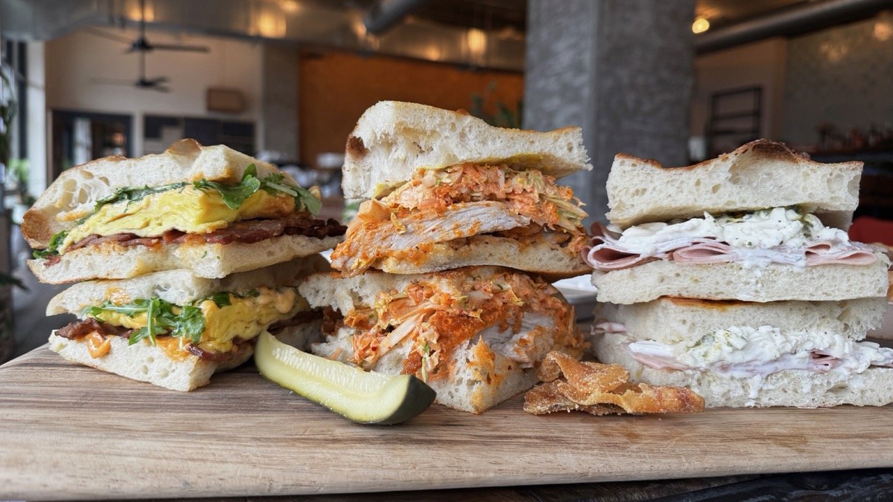Three different sandwiches on a wooden serving board, with a pickle spear in front, set in a restaurant or cafe.