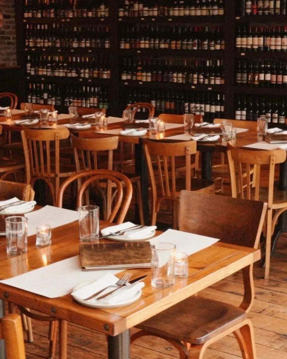 A cozy restaurant interior with wooden tables and chairs, set with white plates, silverware, drinking glasses, and lit candles. A wall of wine bottles lines the background.