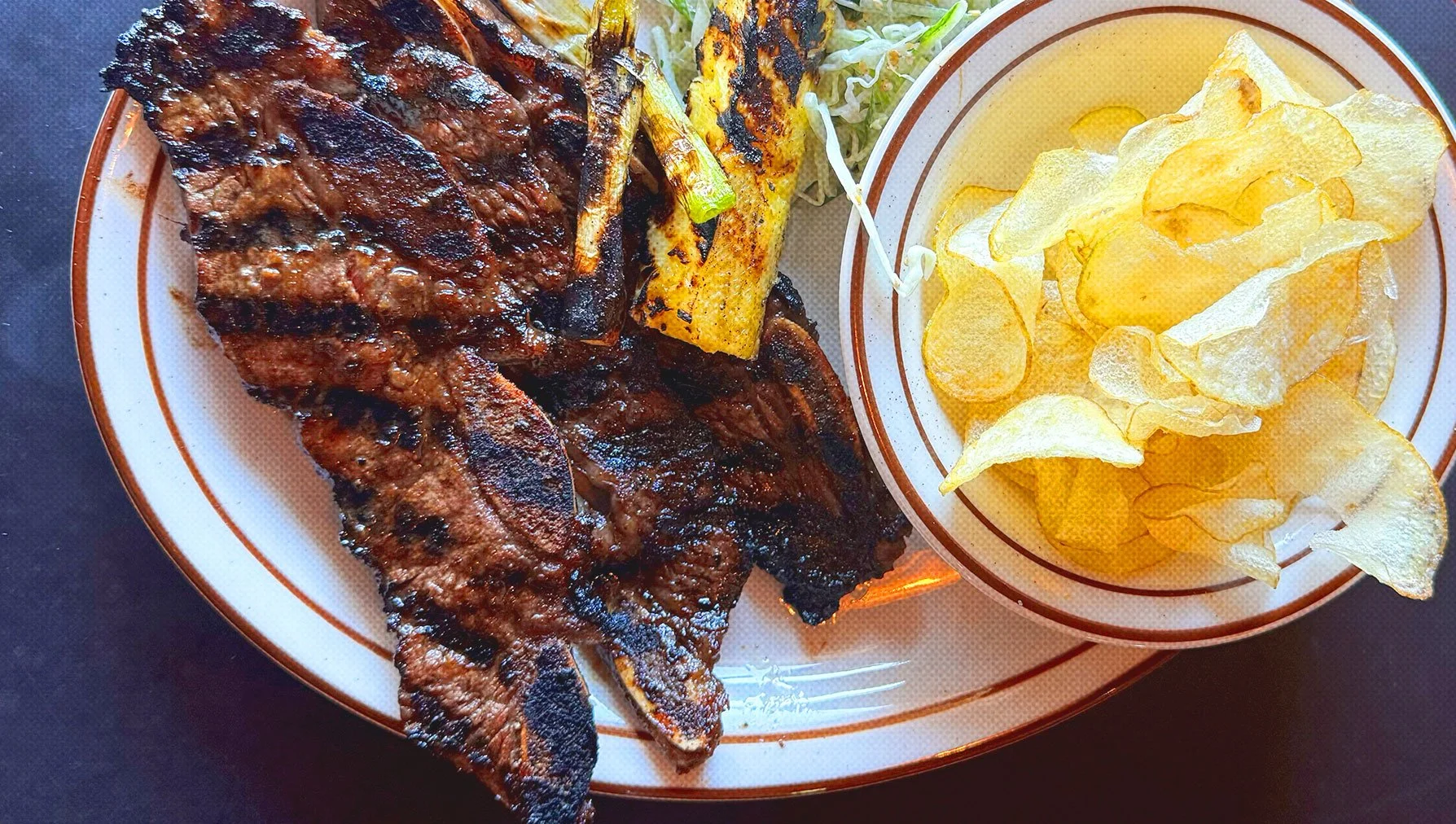 Plate of grilled meat with barbecue sauce, grilled zucchini, a small side of shredded lettuce, and potato chips.