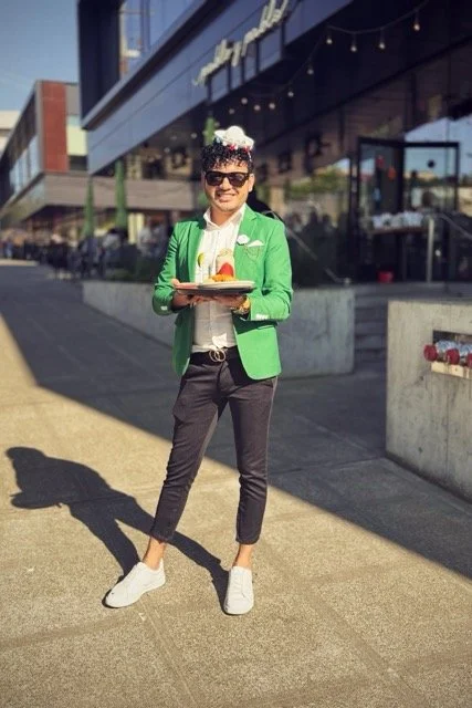 Person standing outside holding a tray of food, wearing sunglasses, a green blazer, and a white shirt, with a festive hat and flowers on their head.