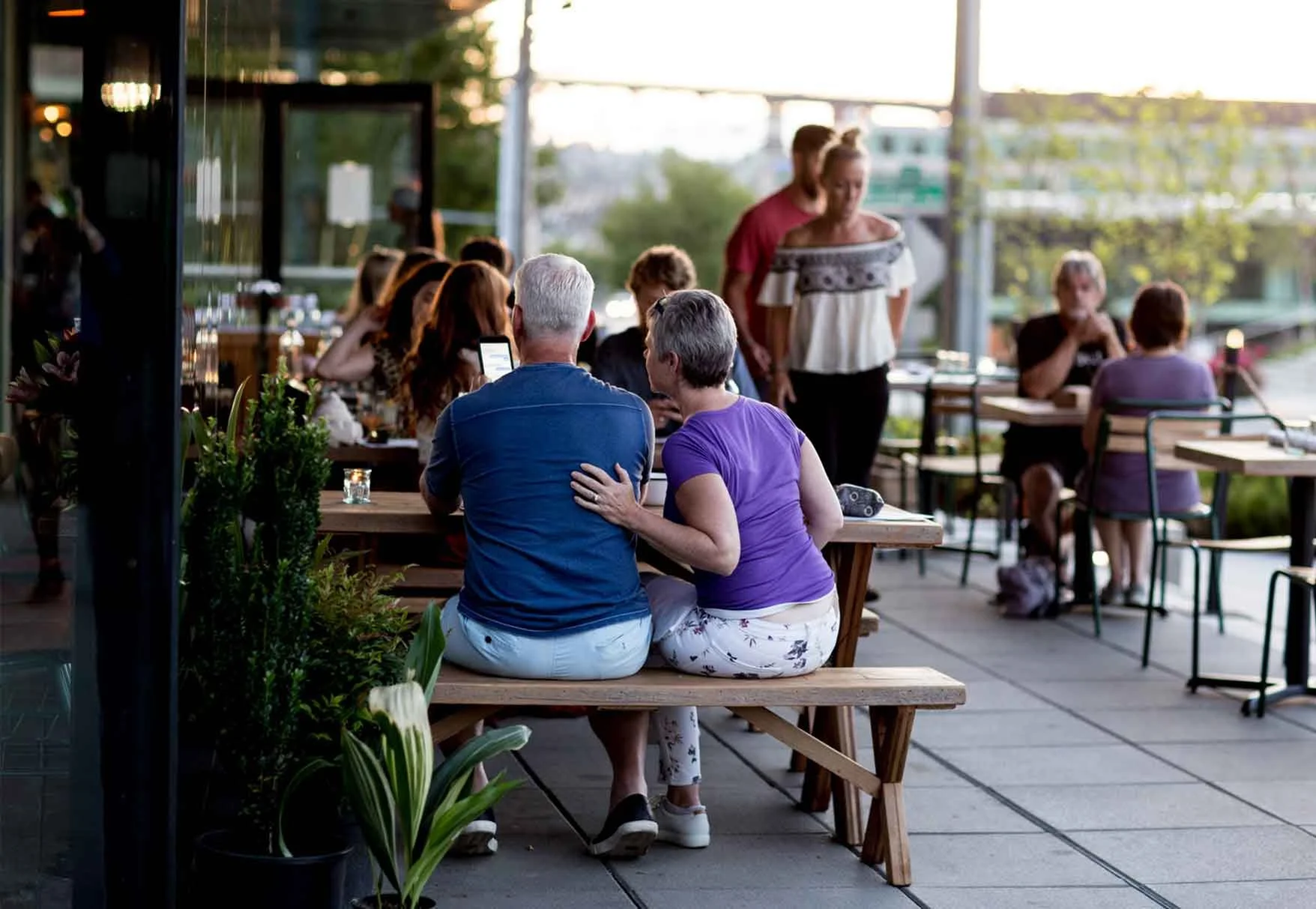 People seated at wooden tables on an outdoor patio at sunset, engaged in conversation, with some using their phones, and a woman with a child standing nearby.