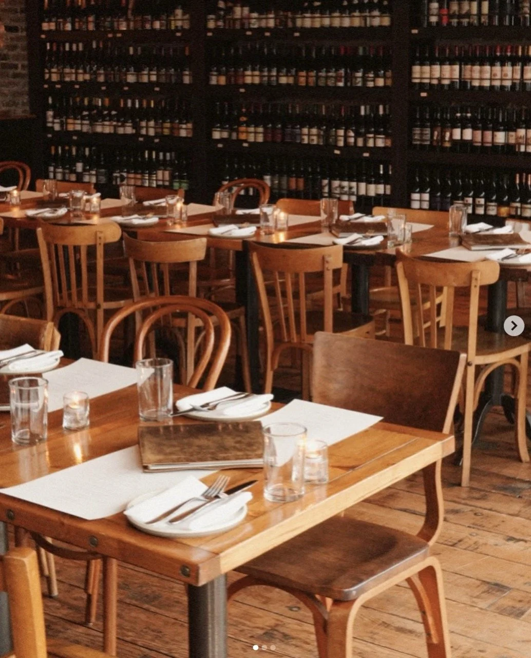 A cozy restaurant dining area with wooden tables and chairs, set with white plates, silverware, glasses, and napkins, and a wall filled with wine bottles.