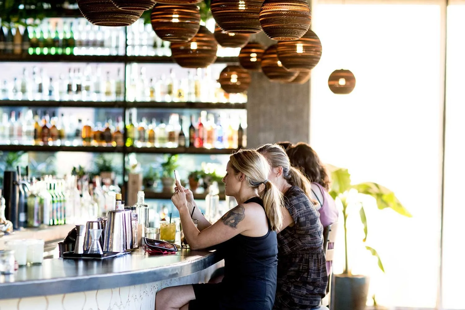 Three women sitting at a bar counter, with one woman taking a photo with her phone and the others sitting beside her, in a well-lit bar with bottles displayed on shelves in the background.