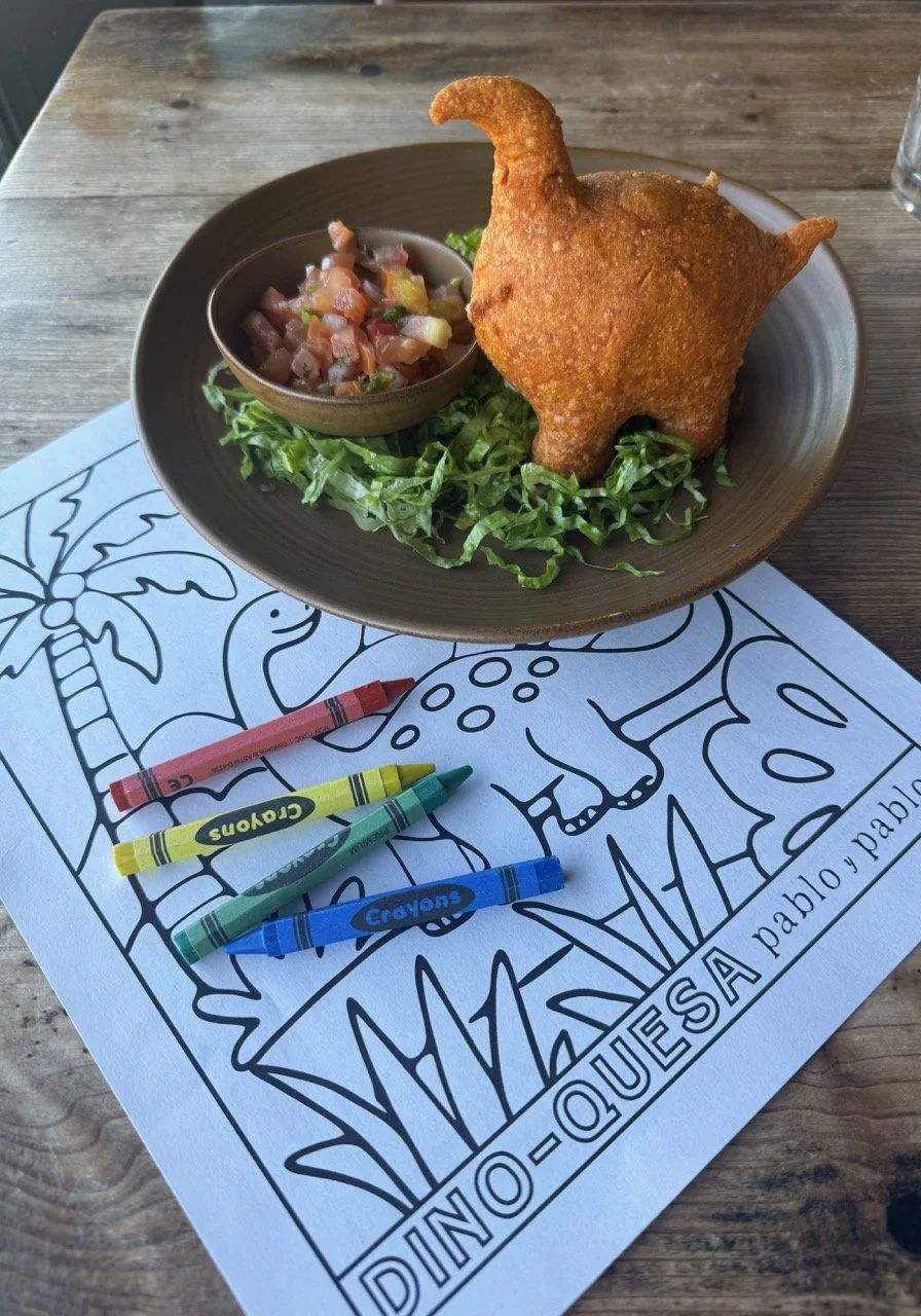 A plate with a deep-fried dino-shaped chicken nugget on a bed of lettuce, served with a small bowl of salsa, on a table with coloring sheets and crayons.