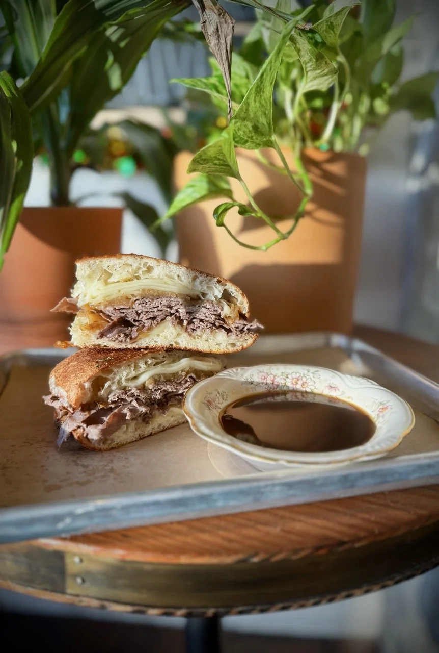 A sliced beef and cheese sandwich on a tray with a small dish of soy sauce or dipping sauce, with potted plants in the background.