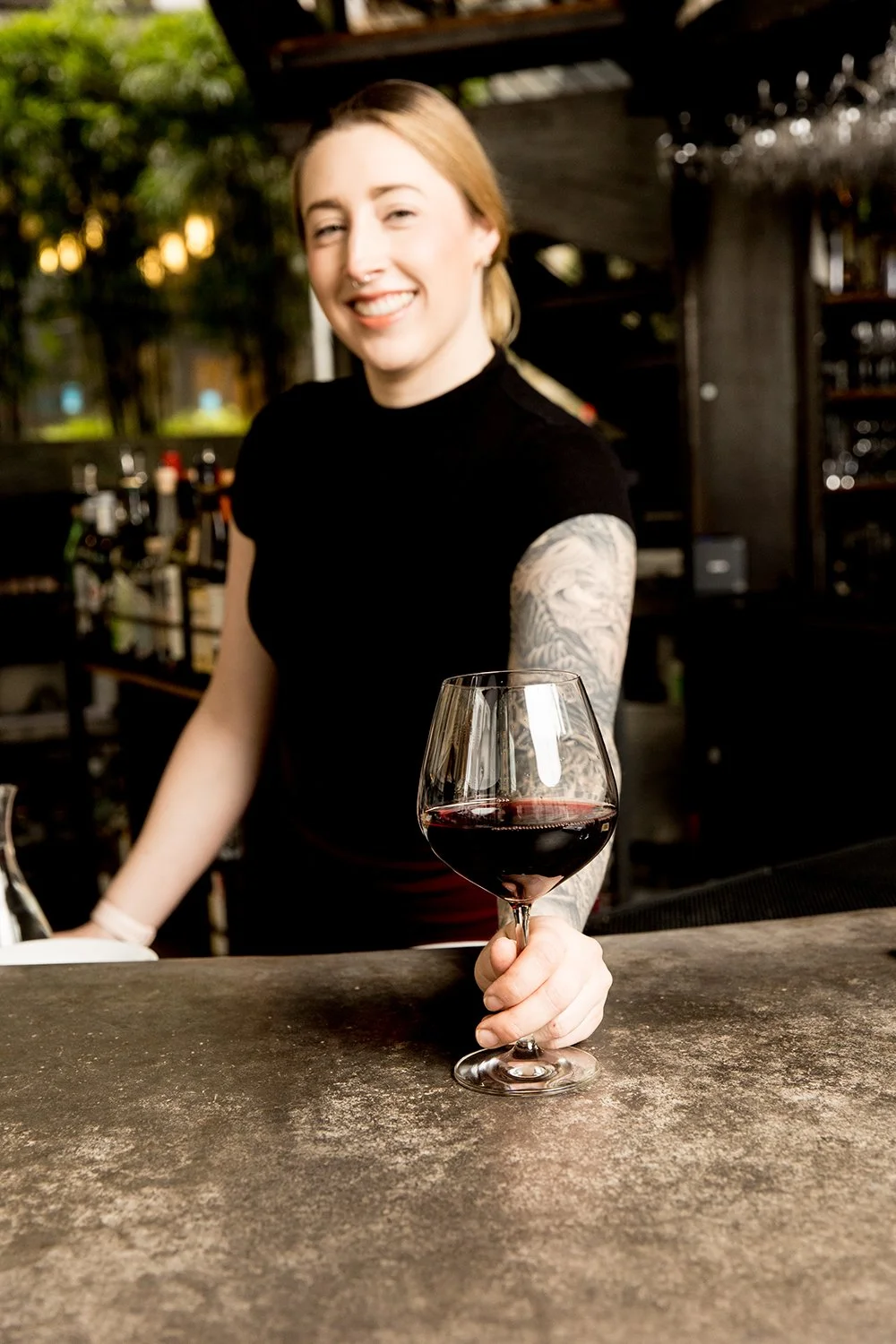 A woman with blonde hair and a tattoo on her arm is smiling and holding a glass of red wine at a bar or restaurant.