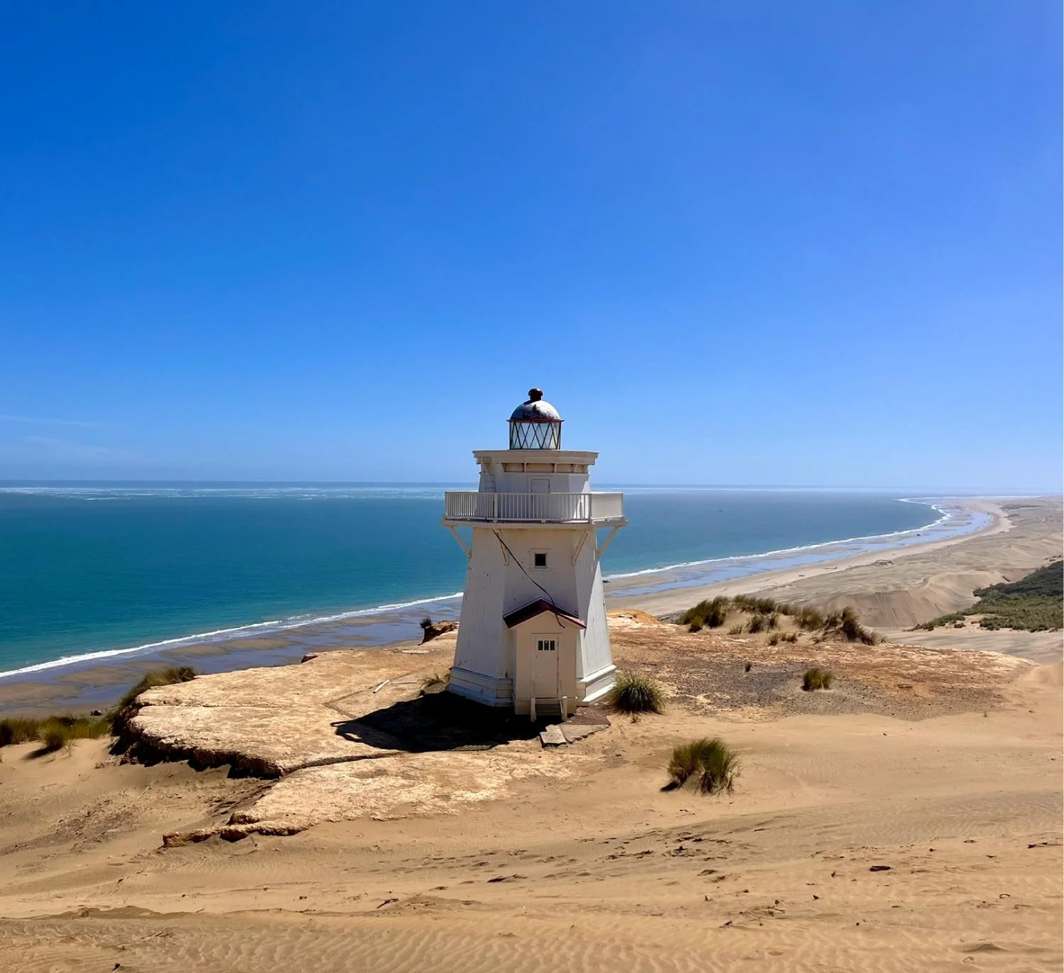 White lighthouse on sandy dunes overlooking the ocean under a clear blue sky.