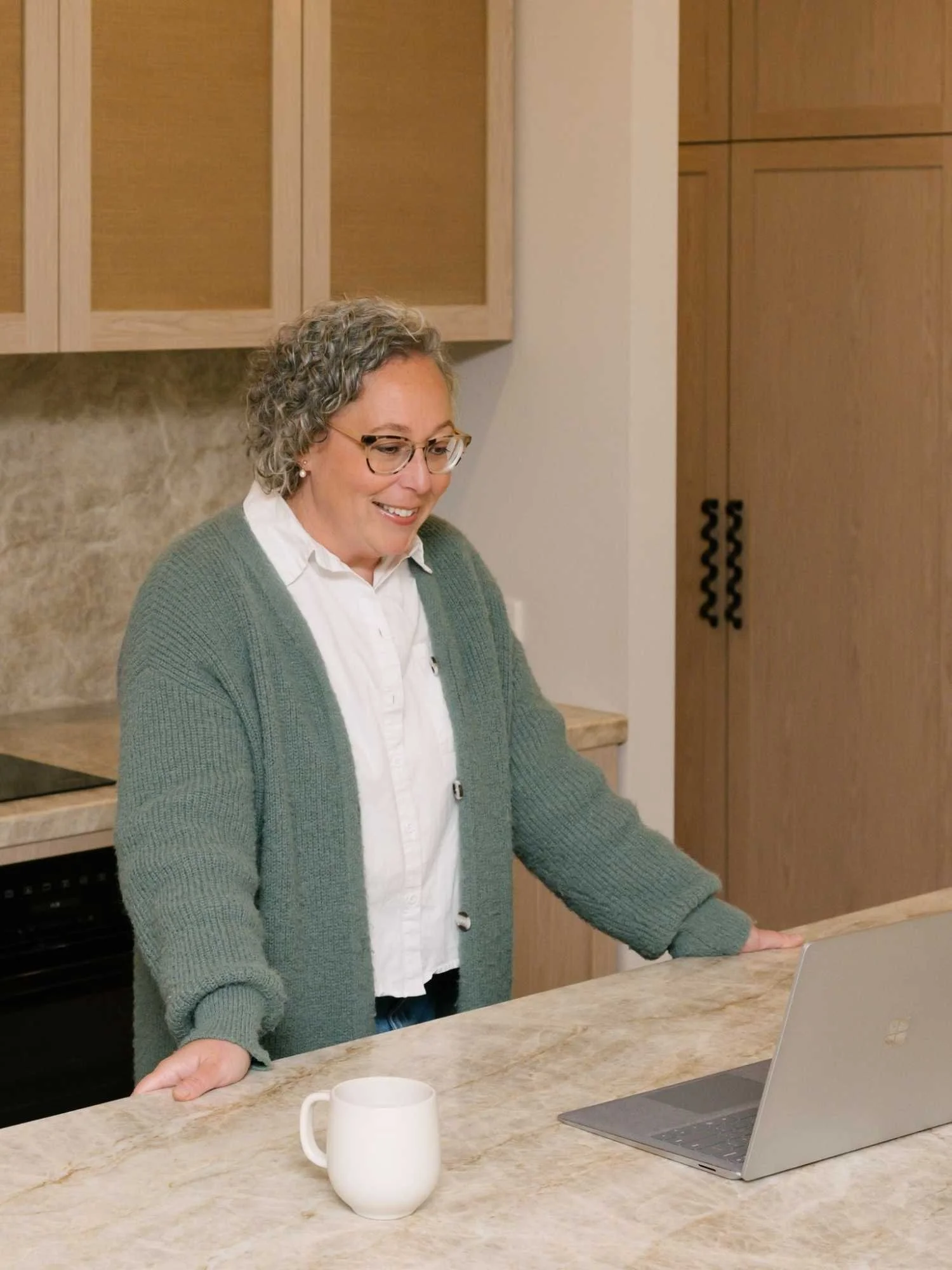 A woman with gray curly hair and glasses, smiling, standing at a kitchen counter with a laptop, looking down, with a white mug in front of her.