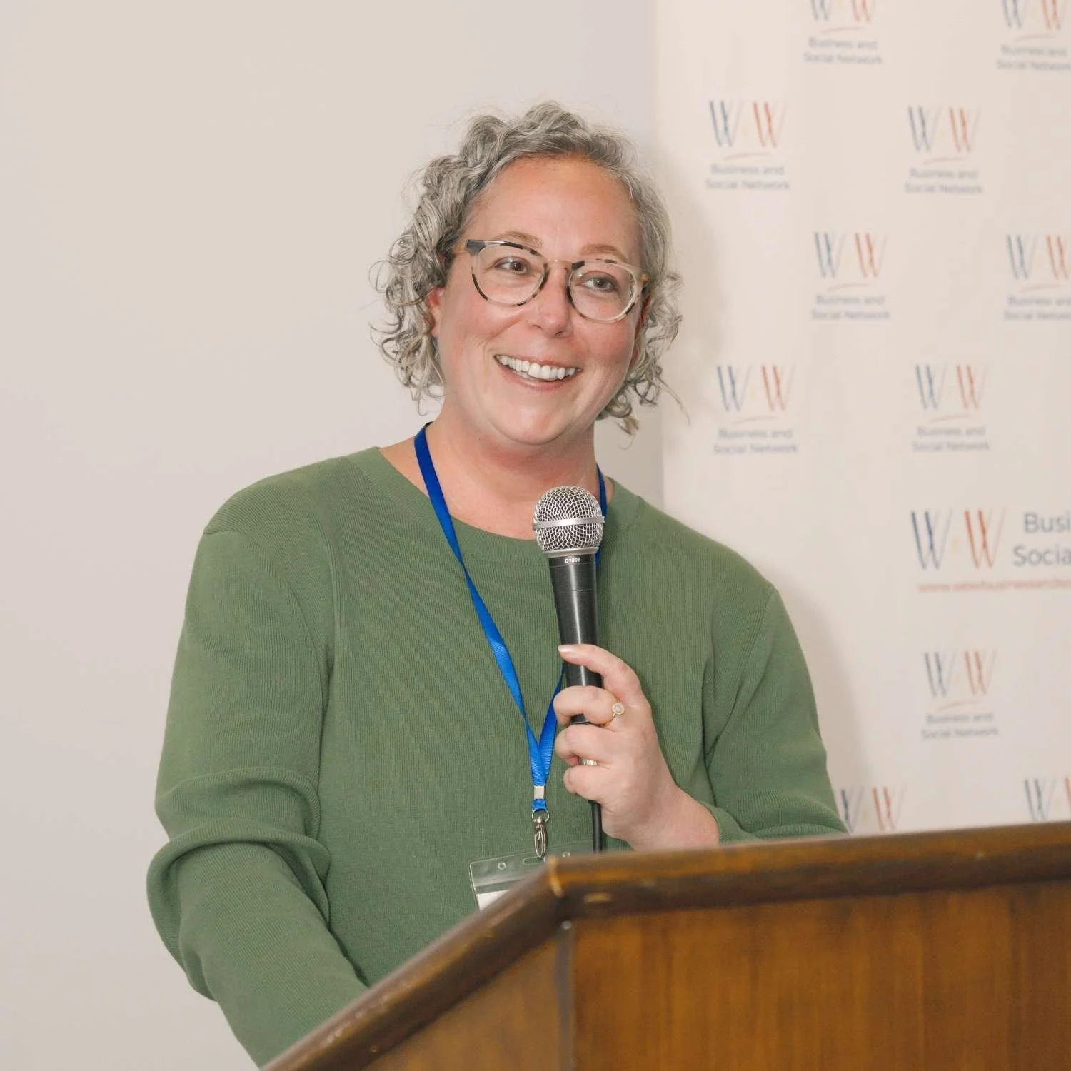 A woman with curly gray hair and glasses smiling while holding a microphone, standing at a wooden podium during a presentation or speech at an event with a branded backdrop.