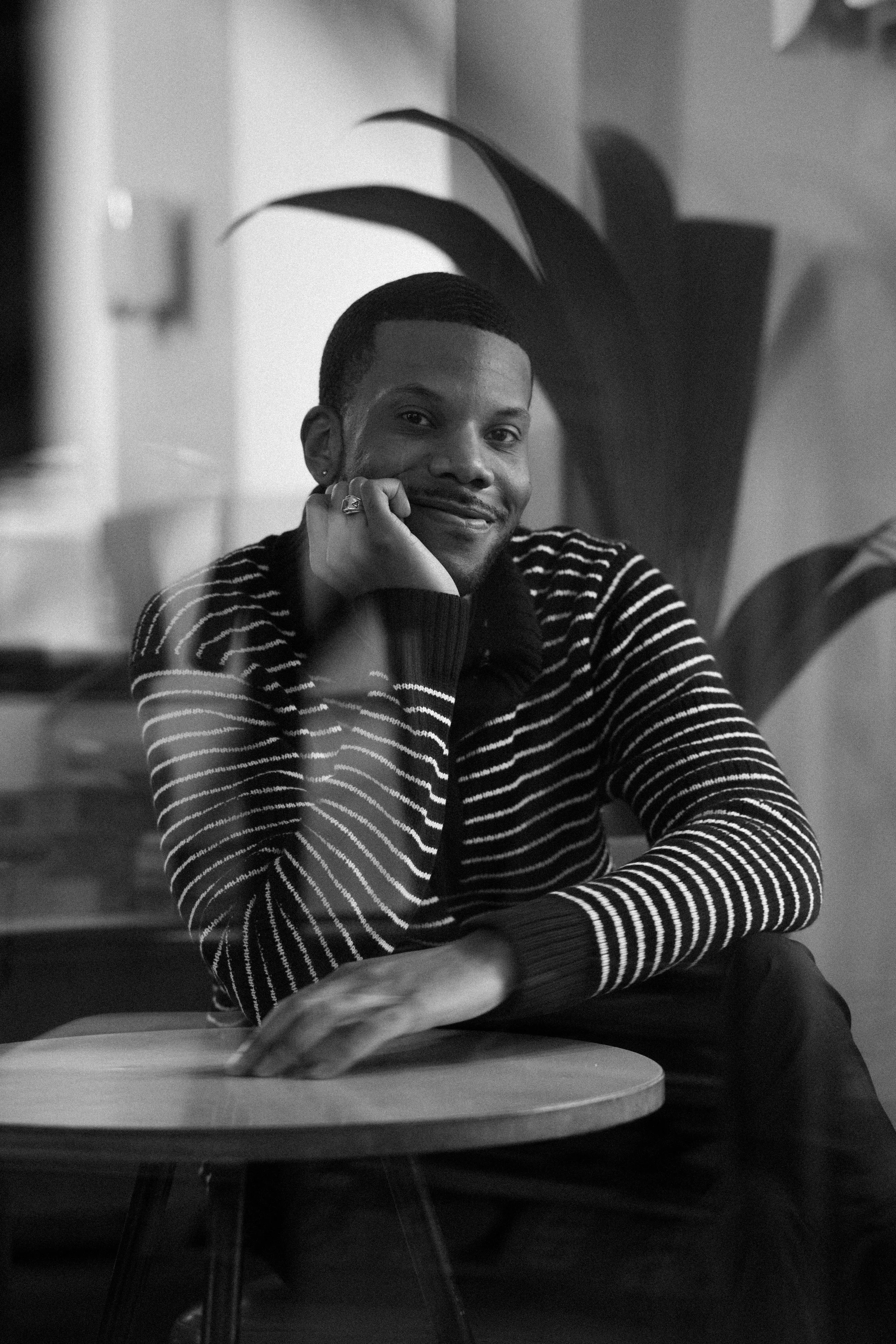 Black and white photo of a smiling person sitting at a table with their head resting on their left hand, wearing a striped sweater, with a large plant in the background.