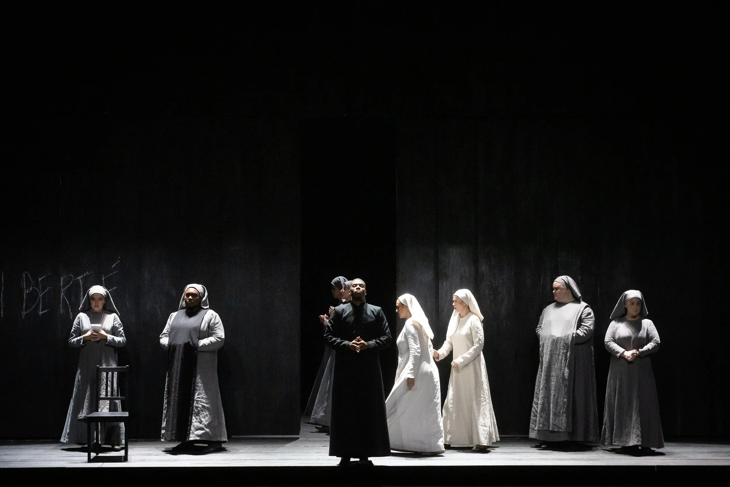 A theatrical performance featuring actors dressed as nuns and monks on a dimly lit stage, with some standing in a line and others engaging in different poses.