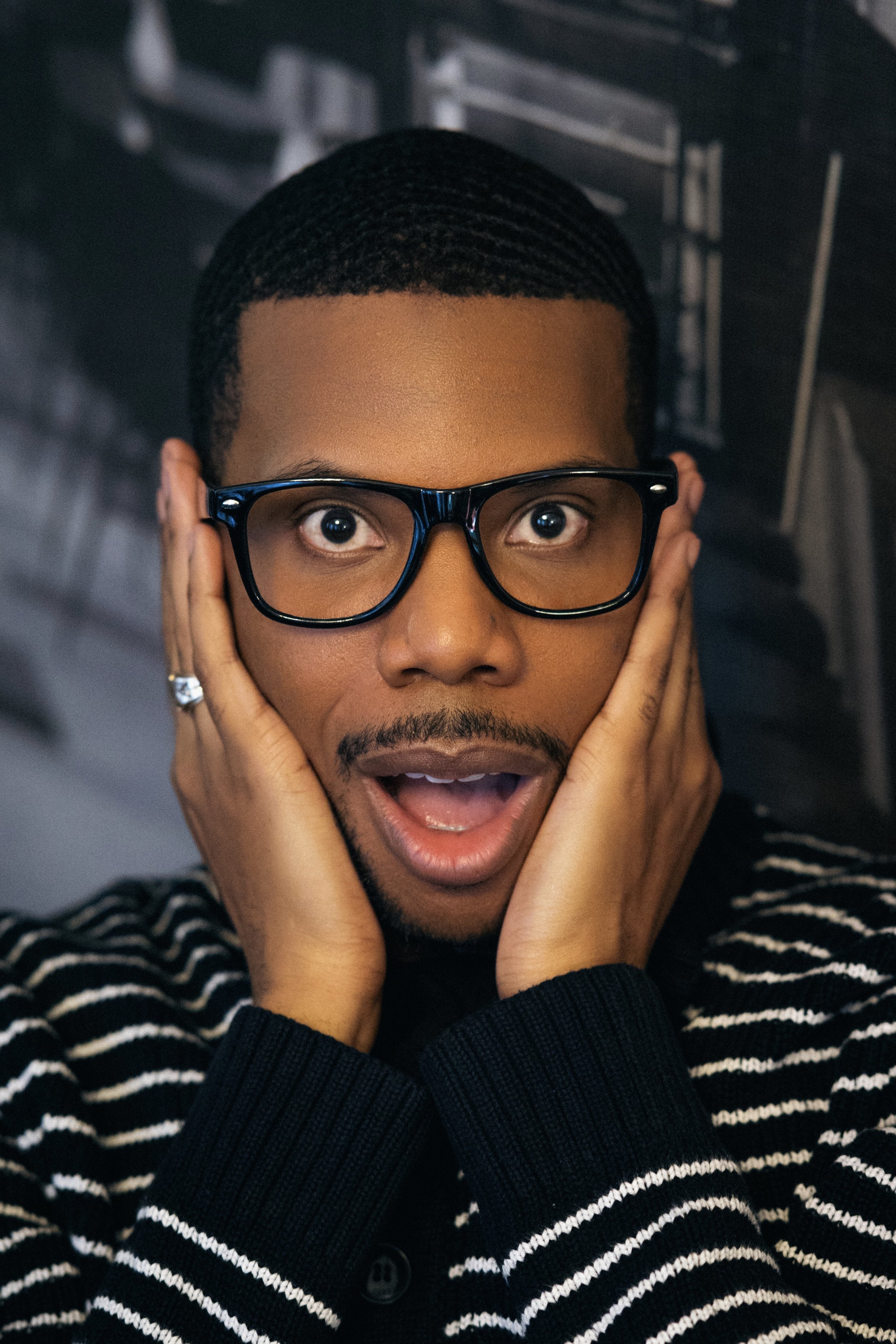 A young man with glasses, surprised expression, hands on his face, wearing a black and white striped sweater.