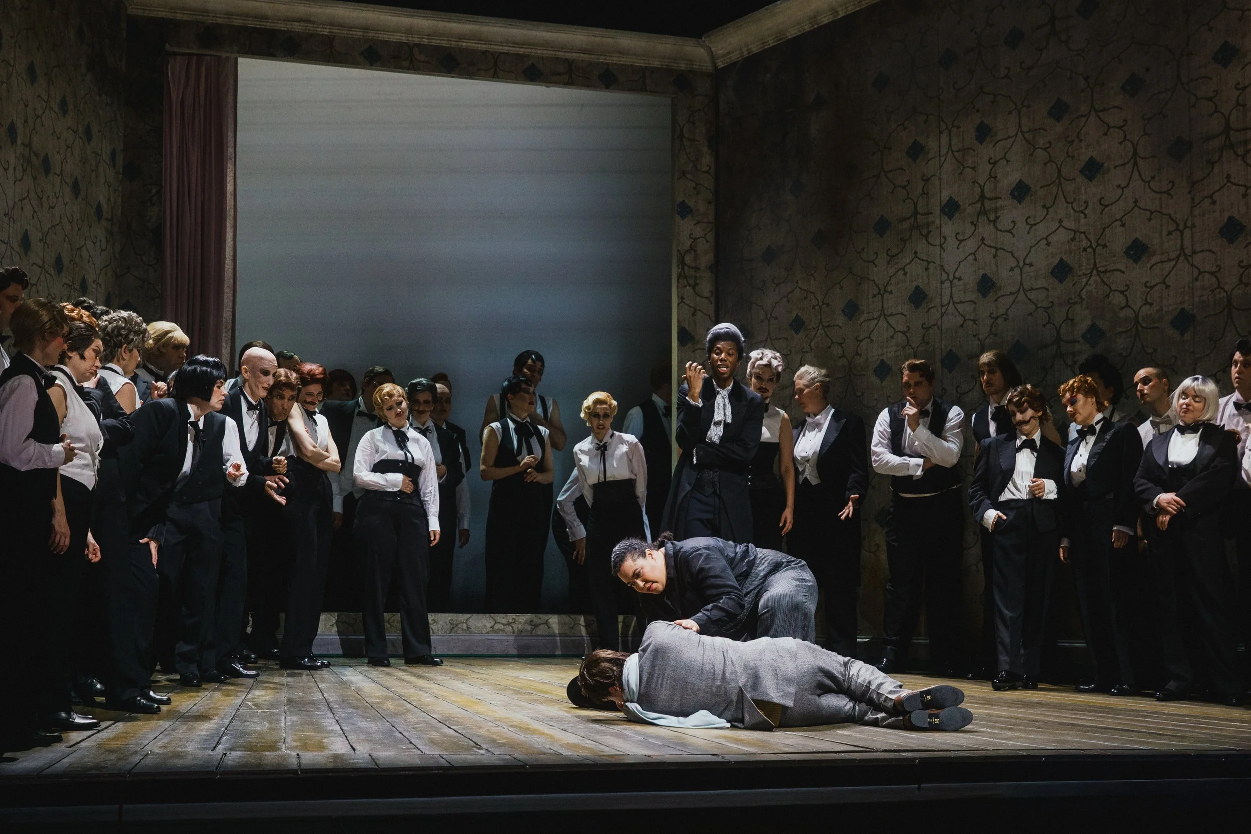 A theatrical performance depicting a group of people dressed in black and white costumes on stage. Two characters are lying on the floor, one handcuffed, with a crowd of onlookers surrounding them. The scene appears tense and dramatic.