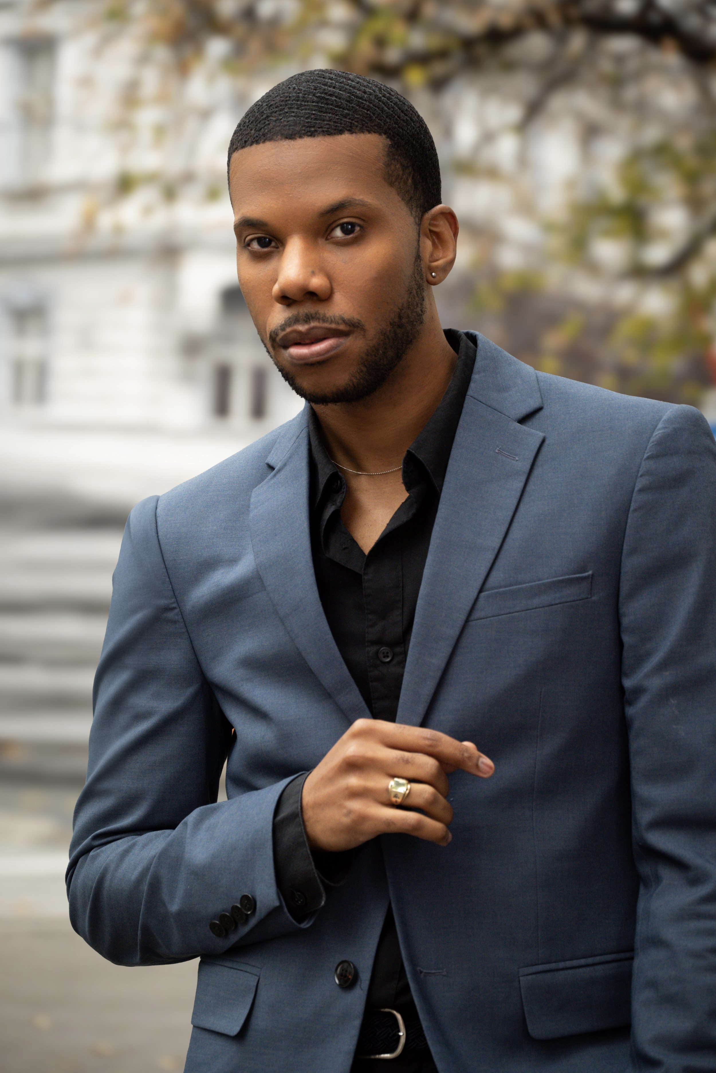Aaron-Casey in a blue suit jacket and black shirt, standing outdoors with blurred trees and building in the background.