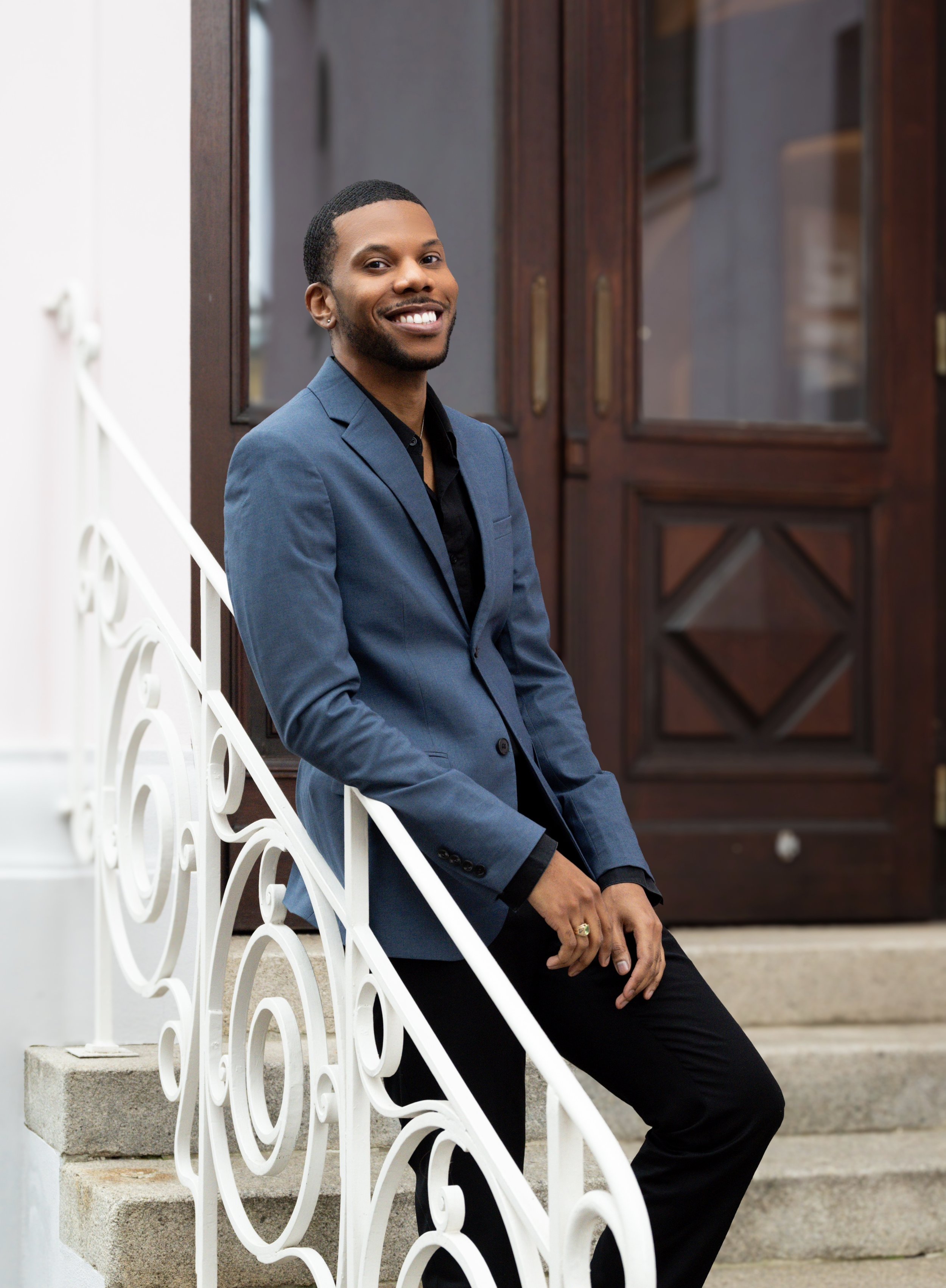 A young man in a blue blazer and black shirt, smiling, sitting on white stairs with an ornate white railing in front of a wooden door with glass panels.