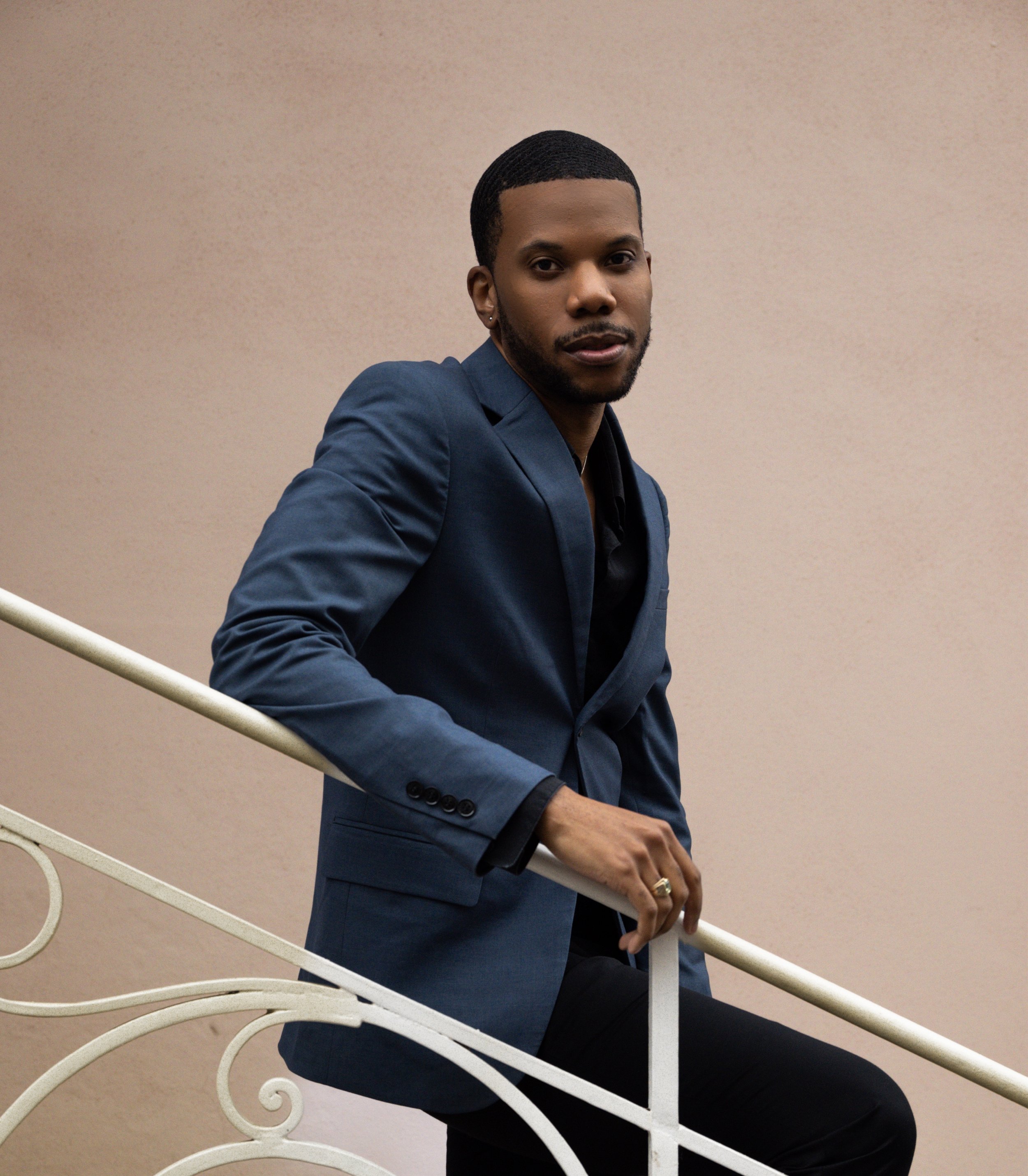 Aaron-Casey dressed in a dark blazer, standing on a staircase with a beige wall in the background.