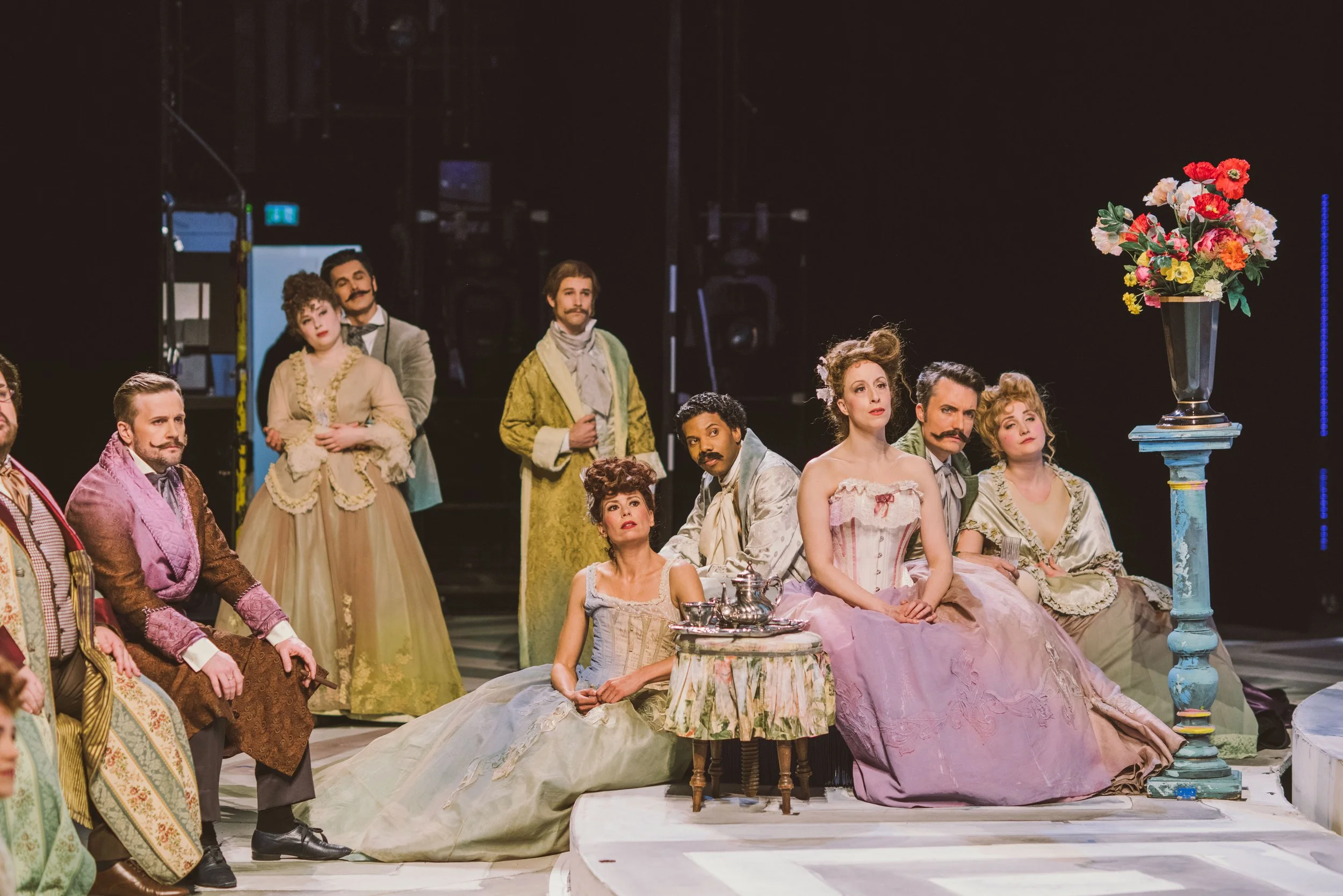 The image shows a theatrical stage scene with a group of actors in historical costumes, seated and standing, with a large vase of colorful flowers placed on a pedestal to the right.