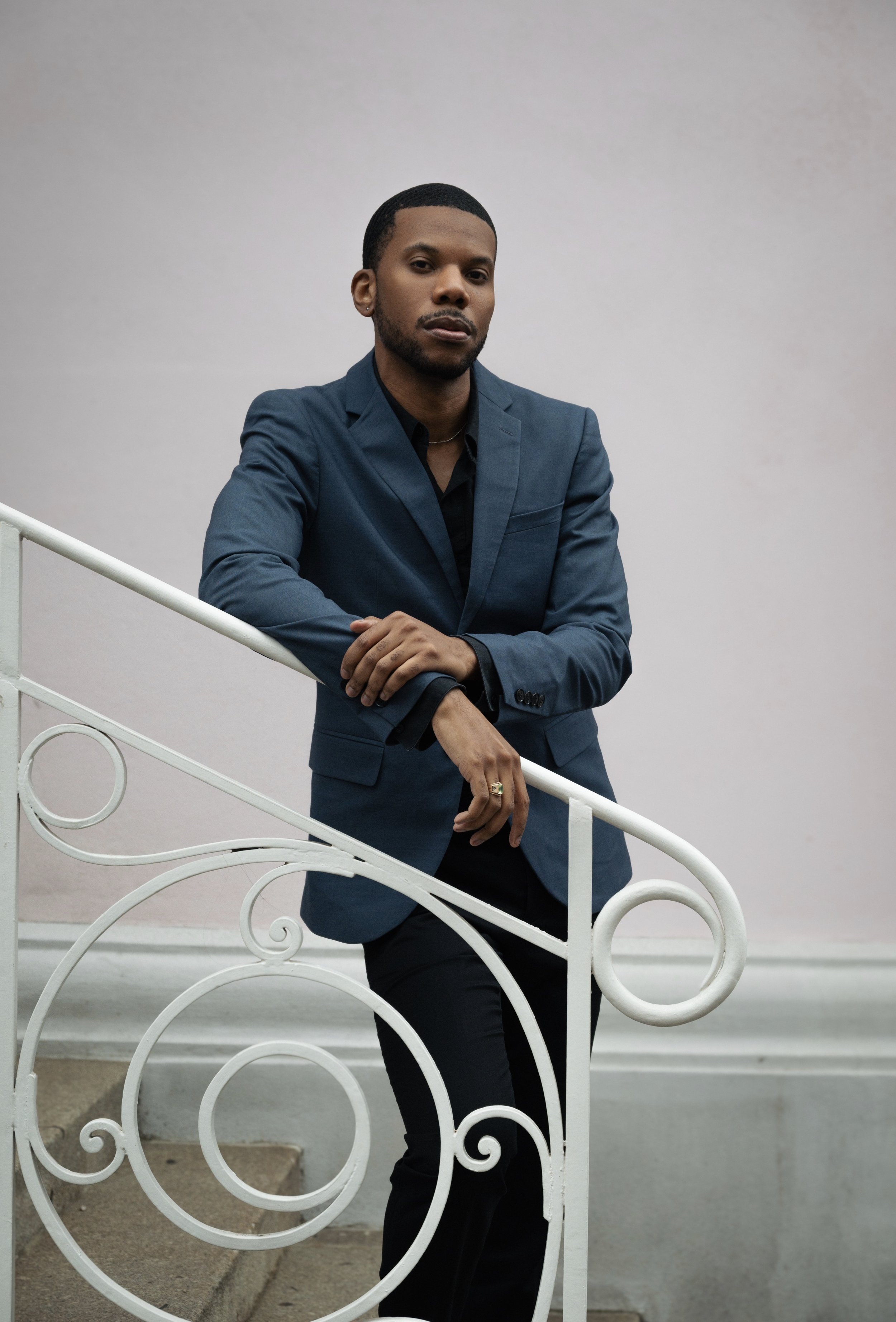 A man in a blue suit and black shirt leaning on a white, ornate metal railing with a white wall behind him.