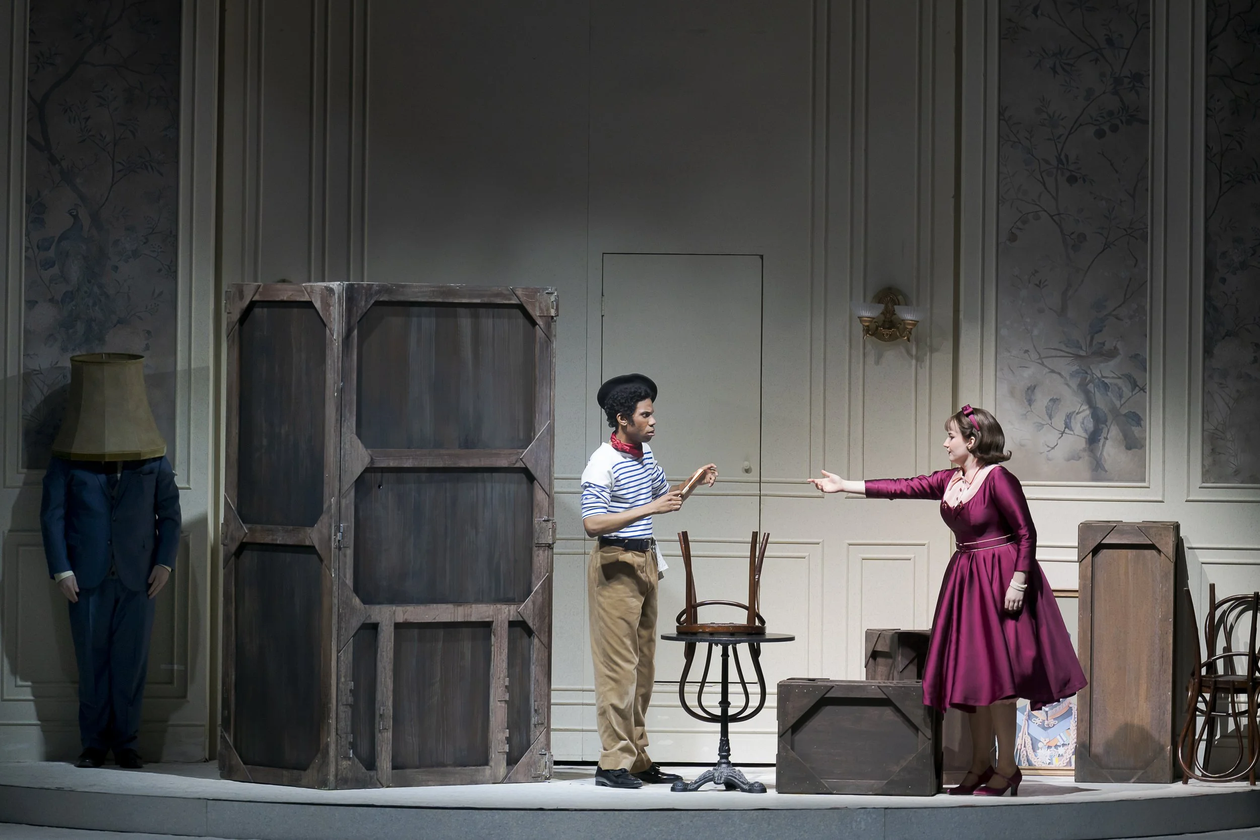 The image shows a theatrical scene with a woman in a red dress reaching out to a woman in a striped shirt, with a man in a dark suit standing with his head bowed. The setting is a decorated room with wallpaper, chairs, and wooden crates.