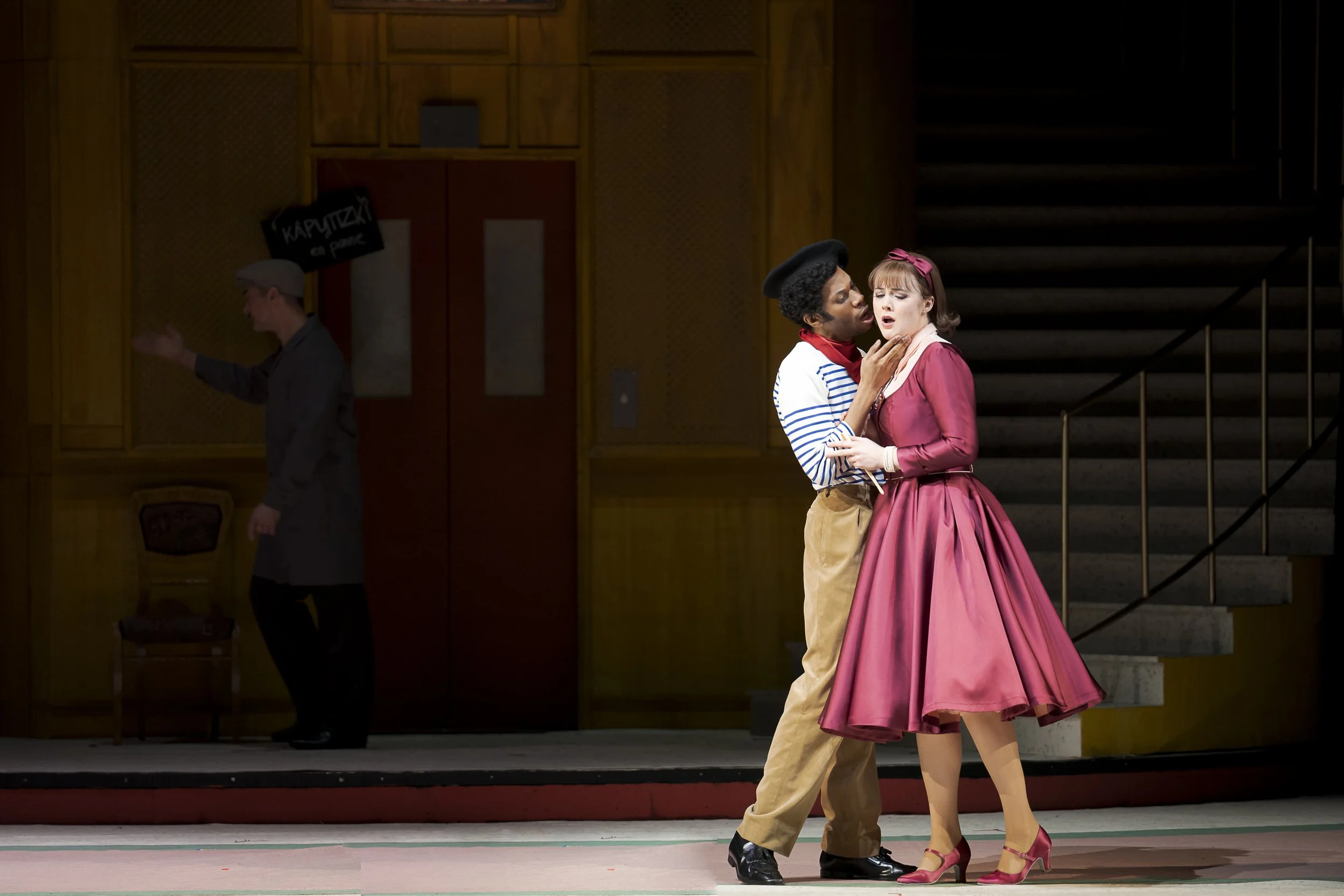 Stage scene with a man and woman performing; the woman wears a pink dress and headband, the man in a striped shirt and beret, with another man in the background near a door.