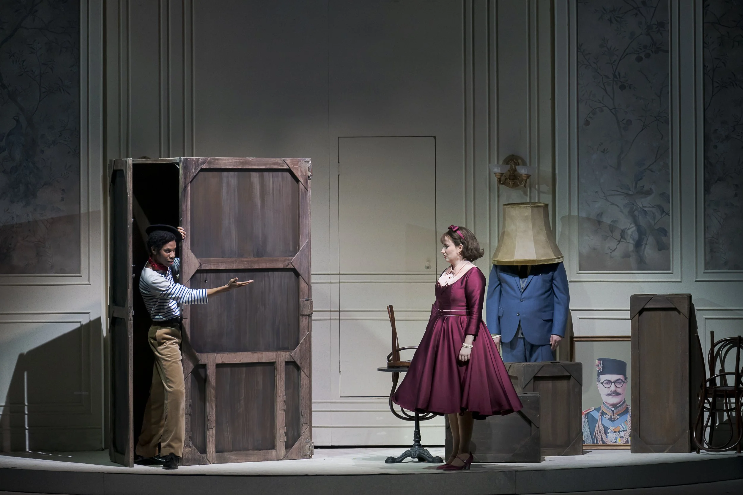Actors performing a scene on stage depicting a woman in a pink dress and a man with a head for a lamp, with other actors and props in the background.