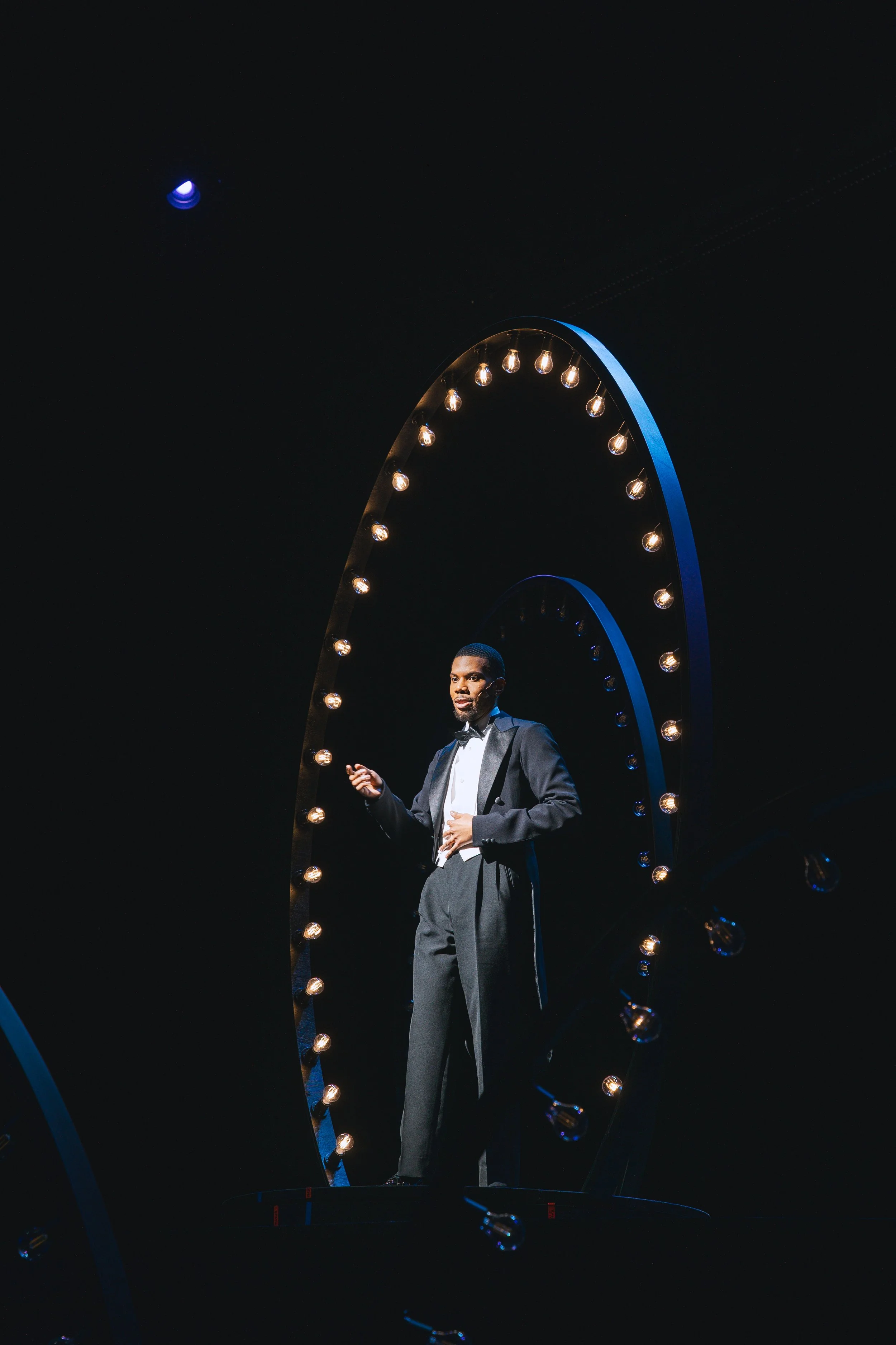 A man in formal attire standing on a stage with a large decorative arch illuminated with small light bulbs.