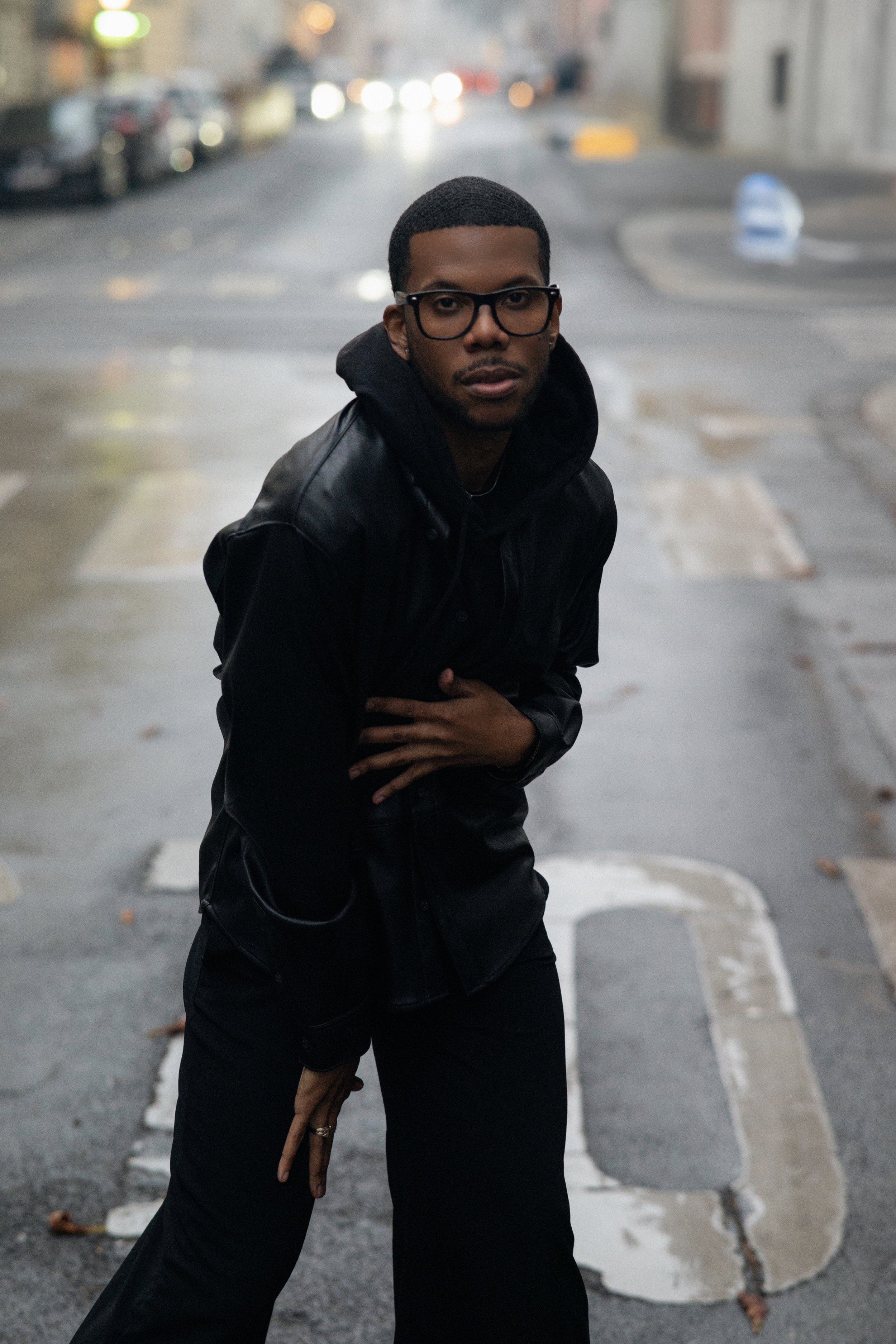Aaron-Casey wearing a black leather jacket and black hoodie, posing outdoors on a city street on a rainy day, with blurry parked cars and streetlights in the background.