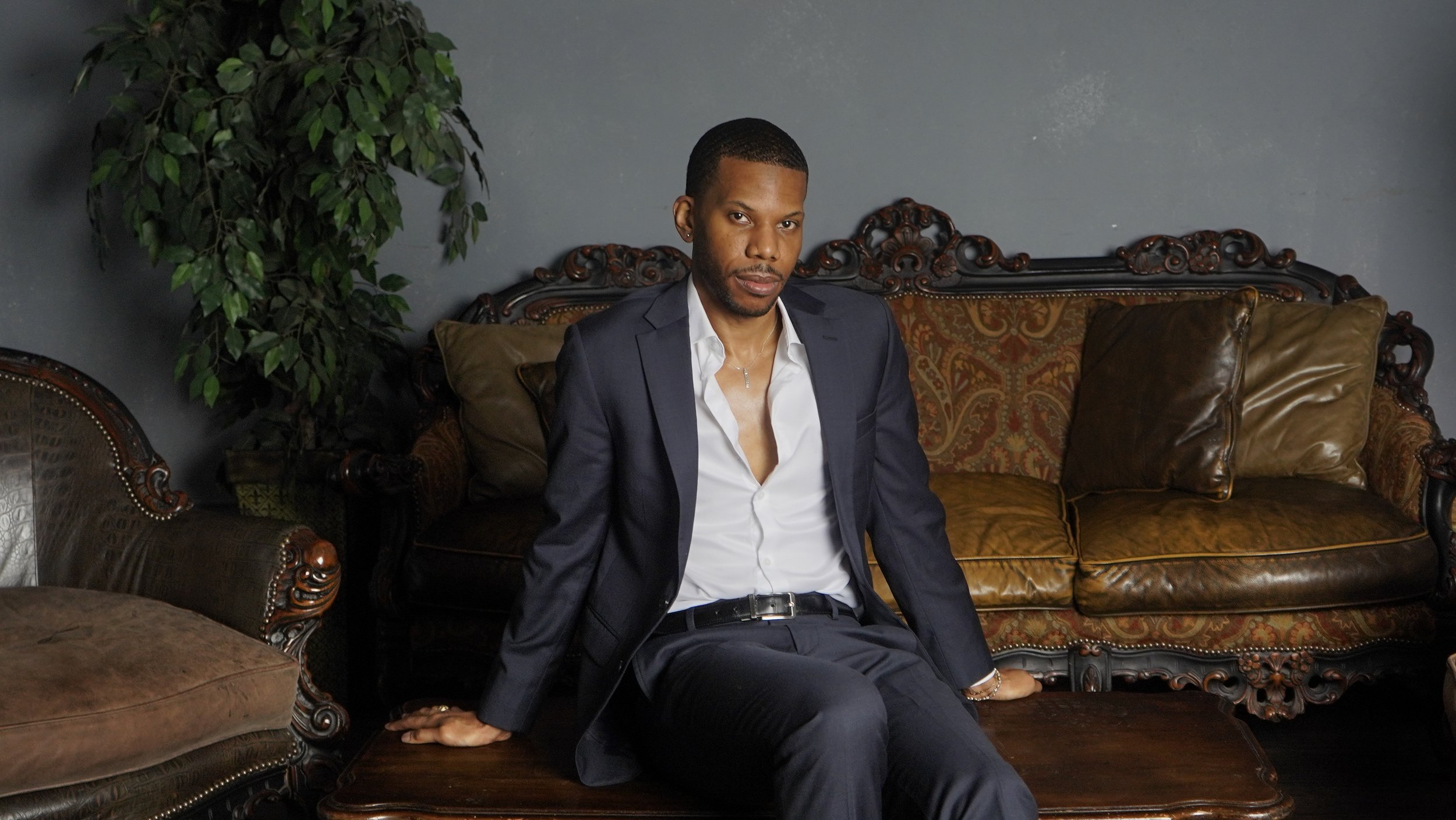 Aaron-Casey in a navy blazer and white shirt sitting on an ornate vintage sofa with brown leather and patterned fabric, with a green plant and a gray wall in the background.