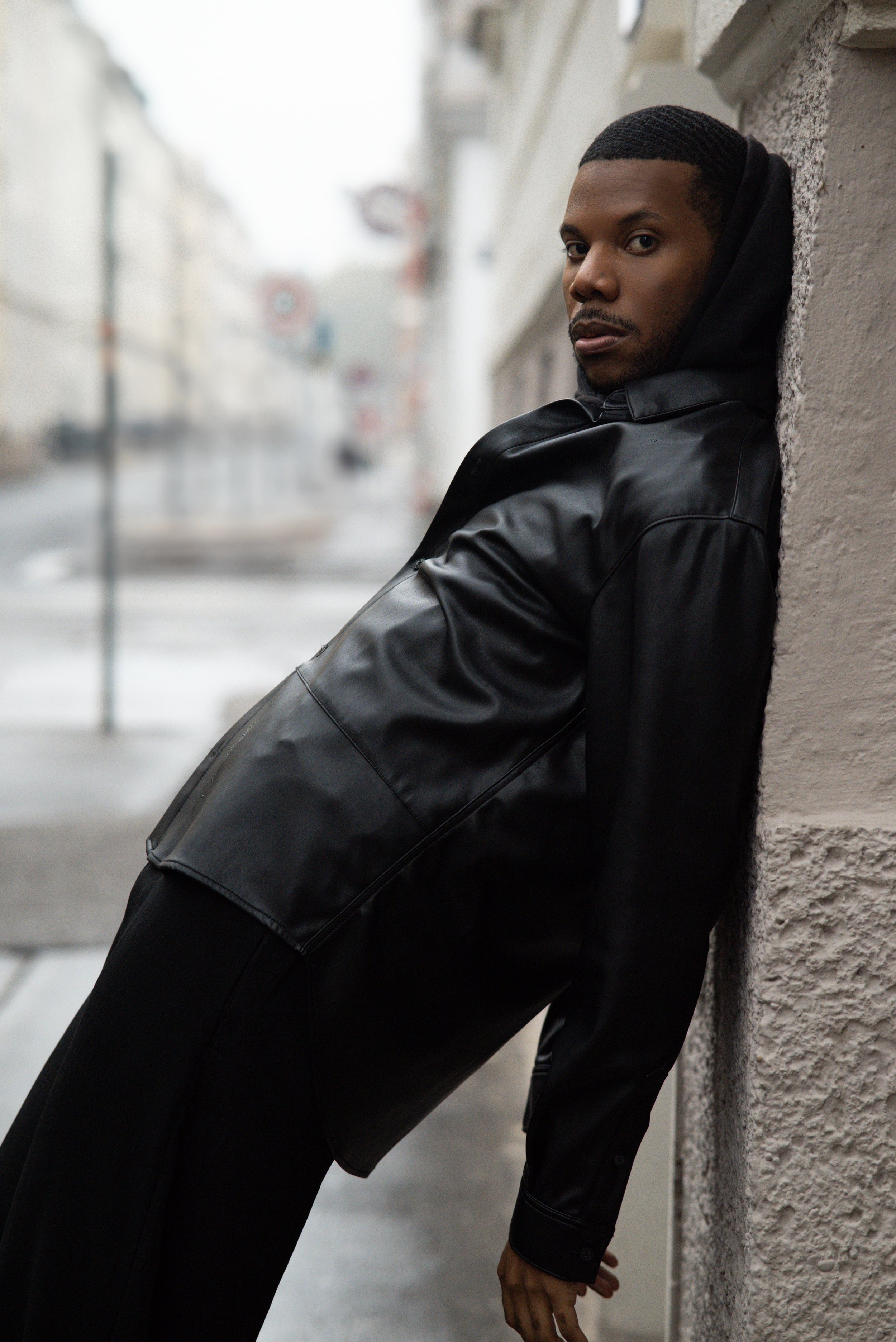 Aaron-Casey wearing a black hoodie and black leather jacket leaning against a textured wall on a city street on a foggy day.