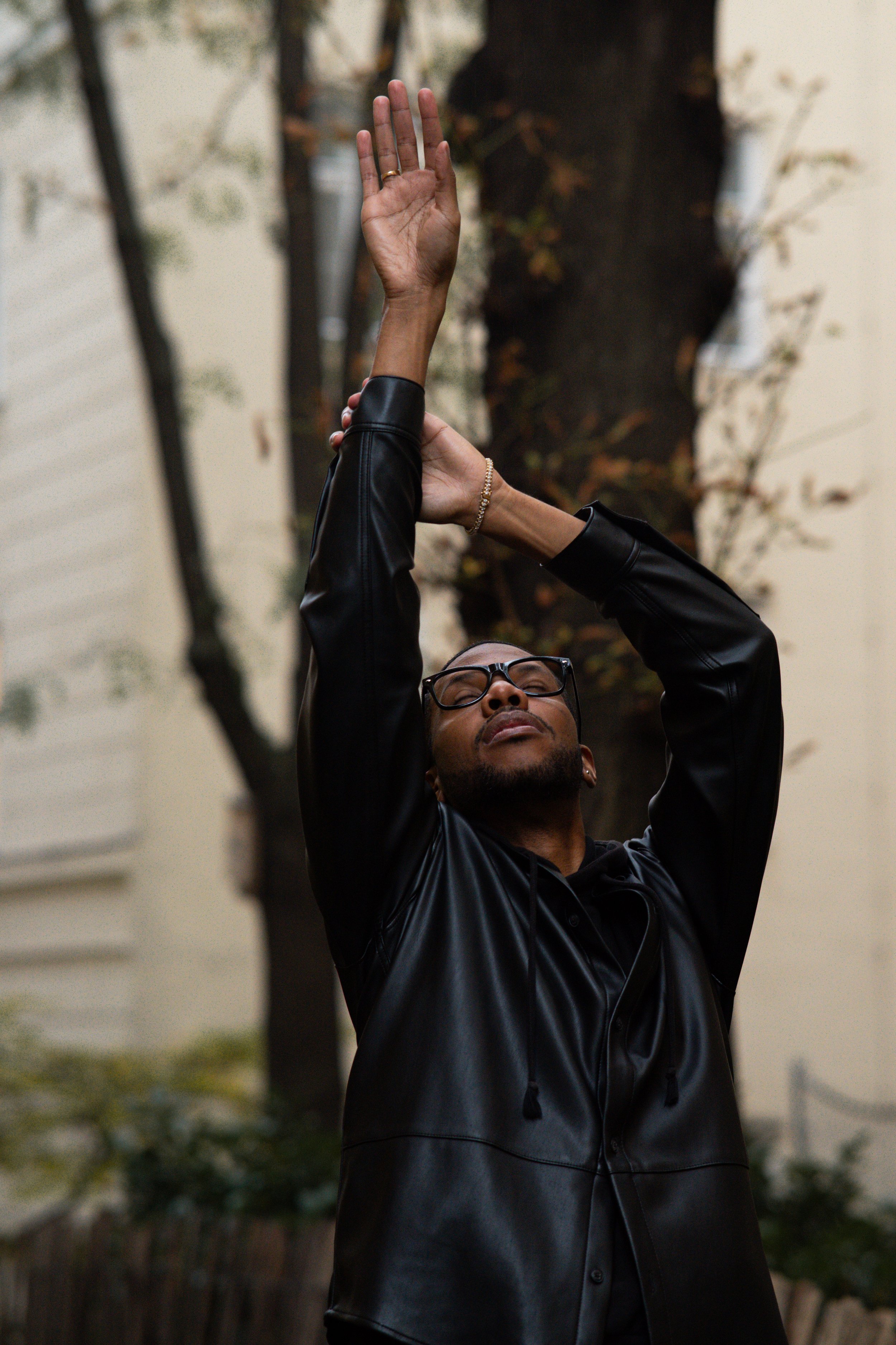 A person with dark skin and glasses wearing a black leather jacket raises a hand with an open palm, with trees and a building in the background.