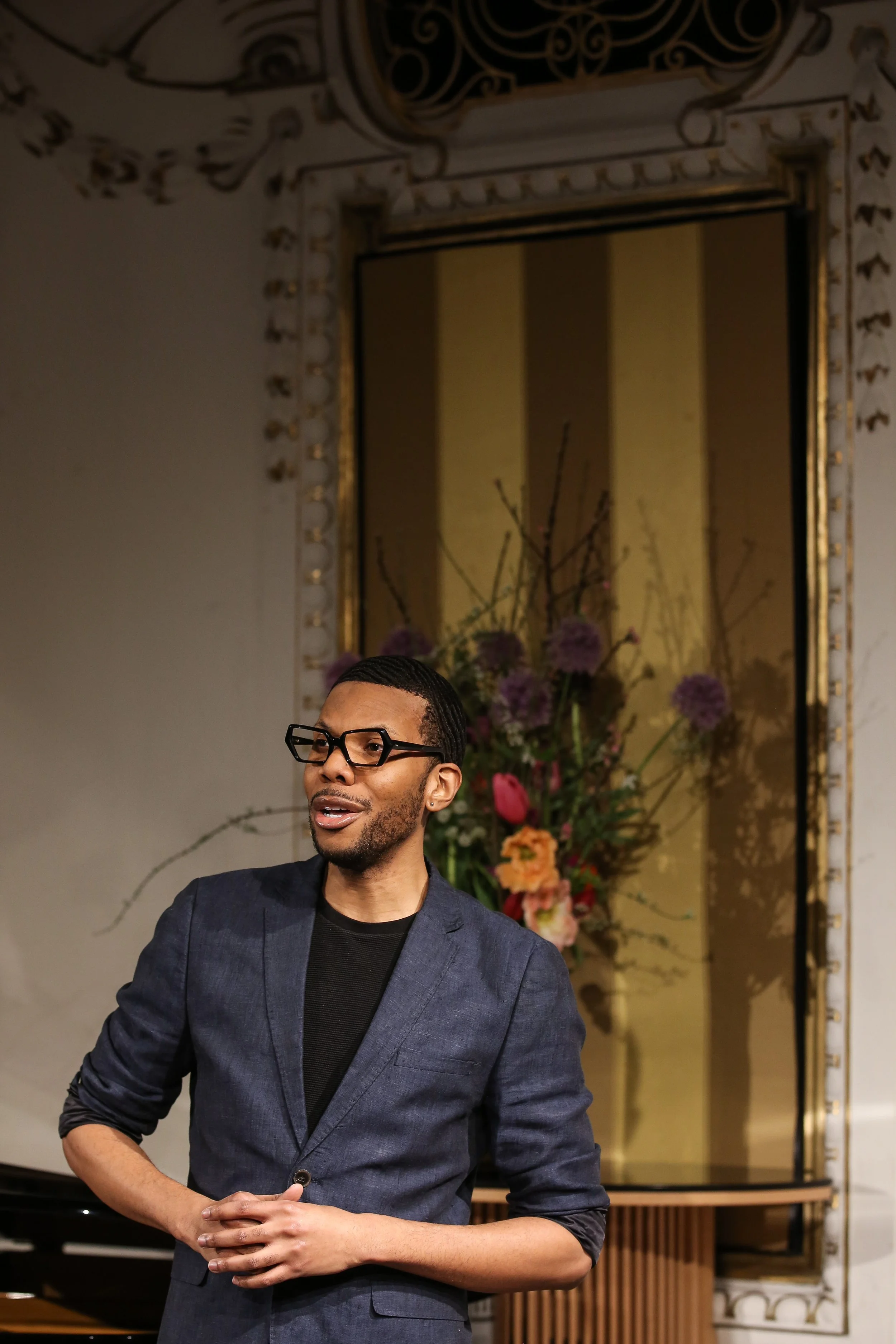 A man wearing glasses and a dark blazer standing in front of a large mirror with ornate gold and white framing, with a floral arrangement of pink, purple, and orange flowers in the background.