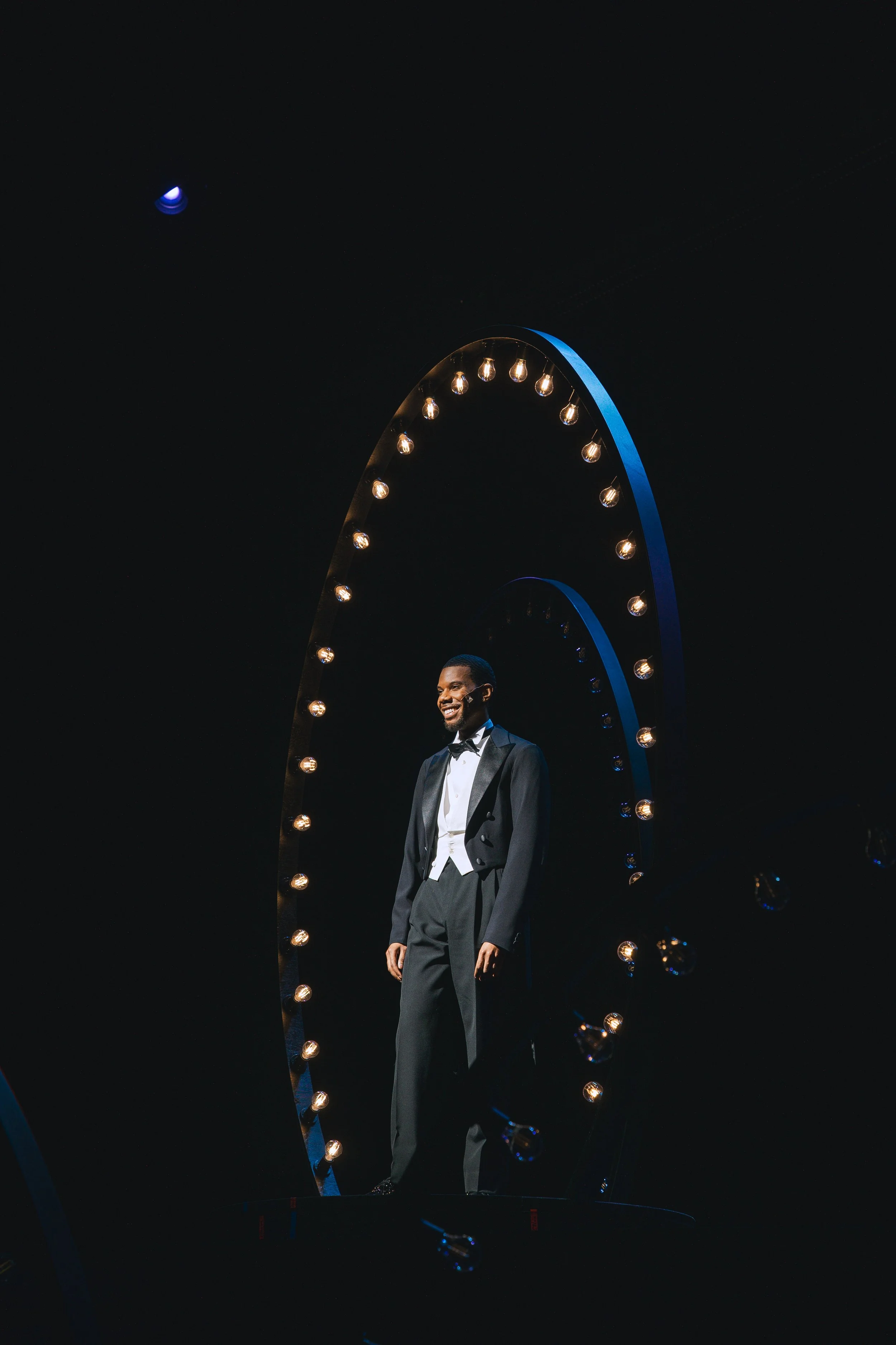 A man in a tuxedo smiling on a stage, standing beneath an illuminated arch at night.