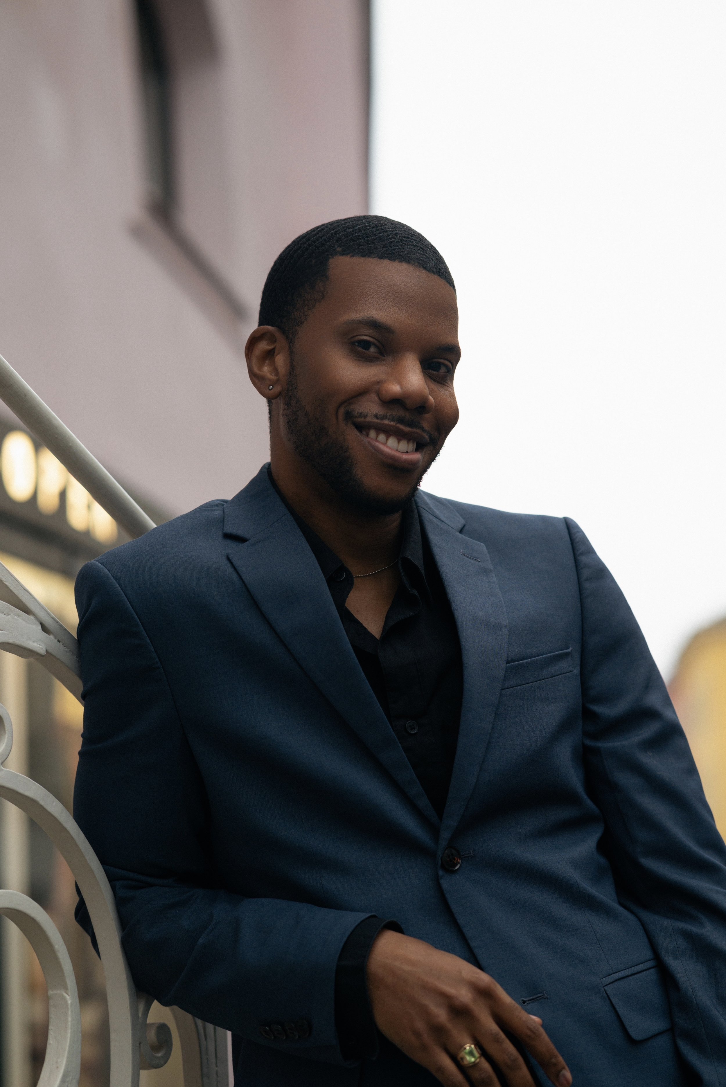 A man dressed in a dark blue suit is smiling and sitting outdoors on a metal staircase, with blurred lights and building in the background.