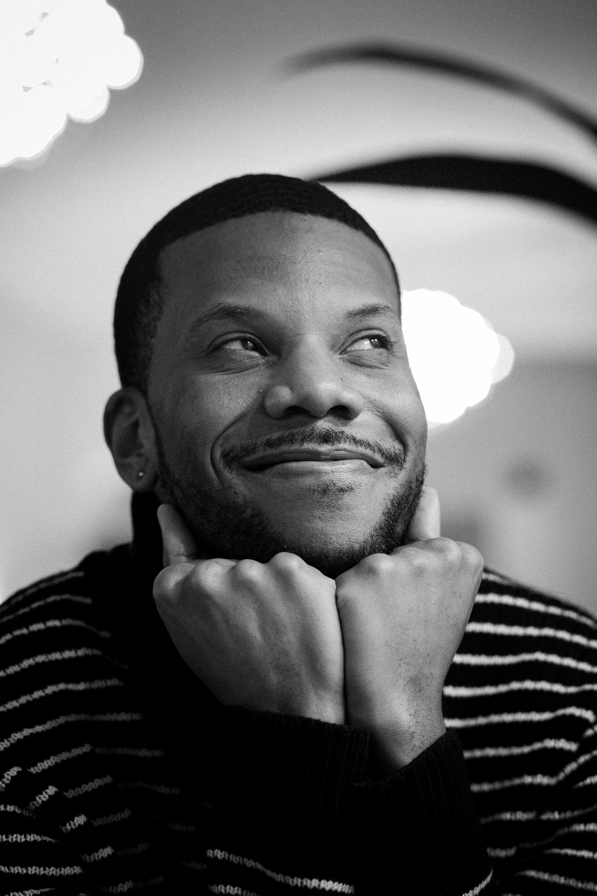 A black and white portrait of a smiling man with short hair, facial hair, and wearing a striped sweater, resting his chin on his hands.