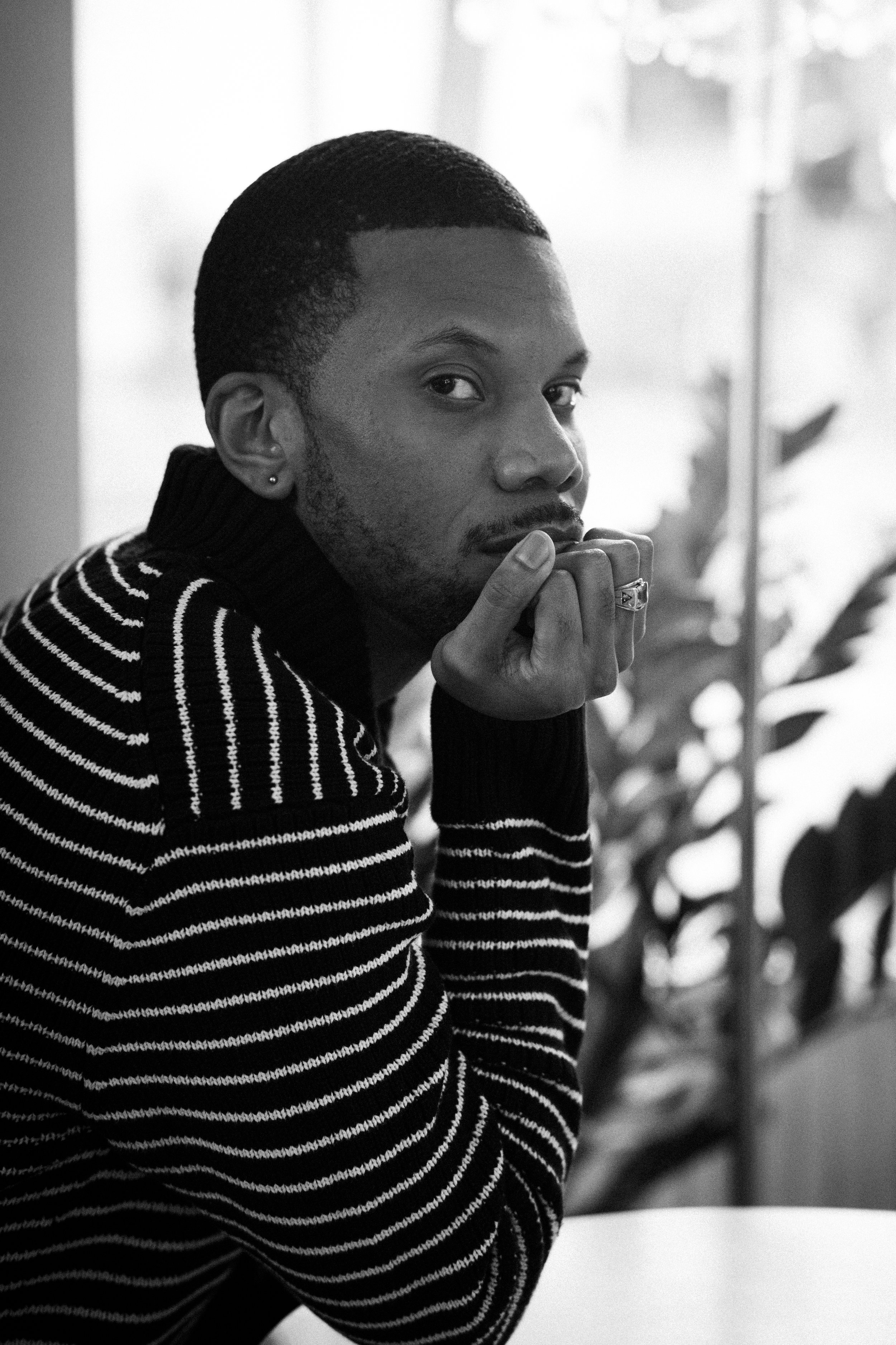 A black and white photo of Aaron-Casey, wearing a striped sweater, resting his chin on his hand with a ring on his finger, looking at the camera.