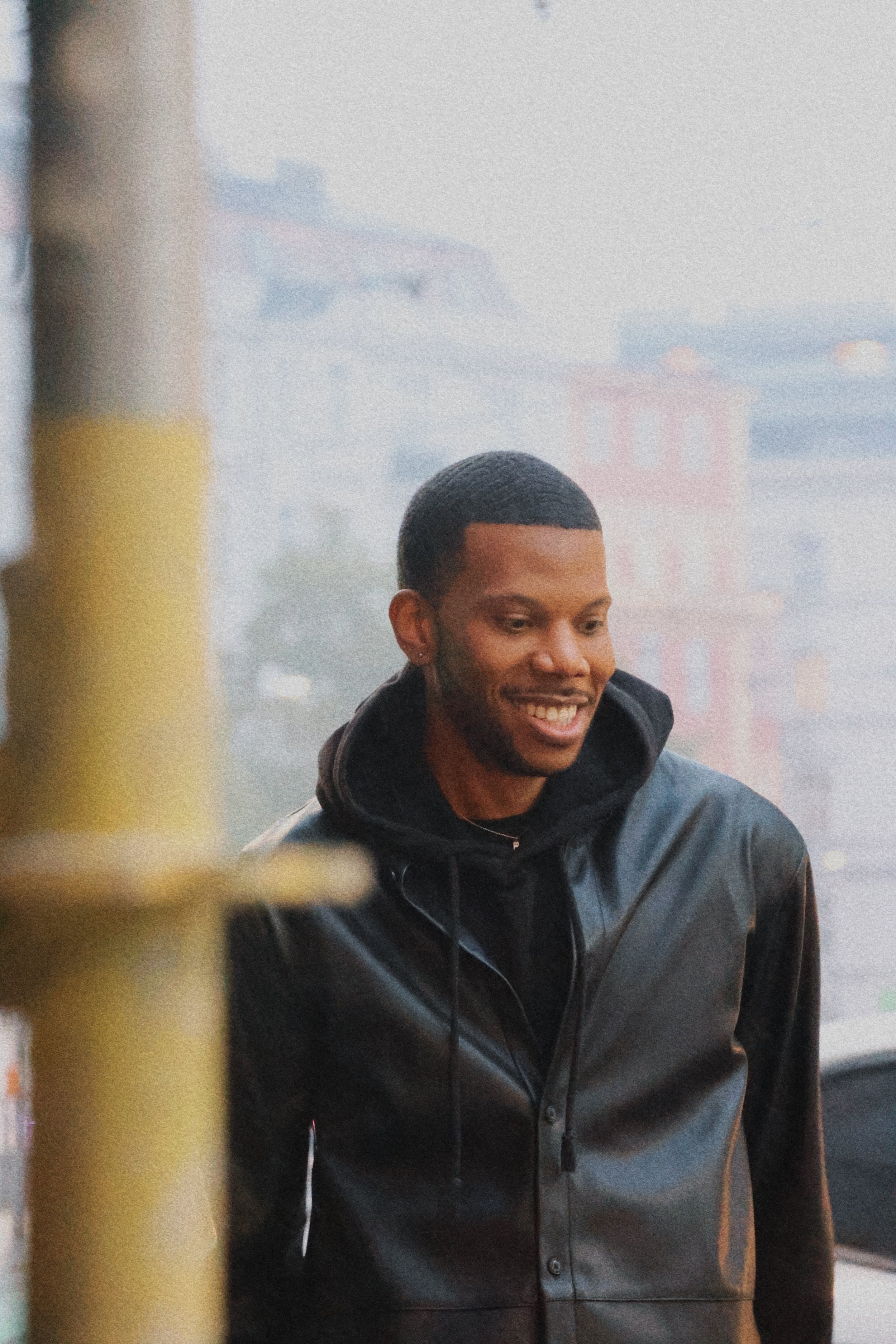 A smiling man with short black hair wearing a black jacket, standing in front of a foggy cityscape with blurry buildings in the background.