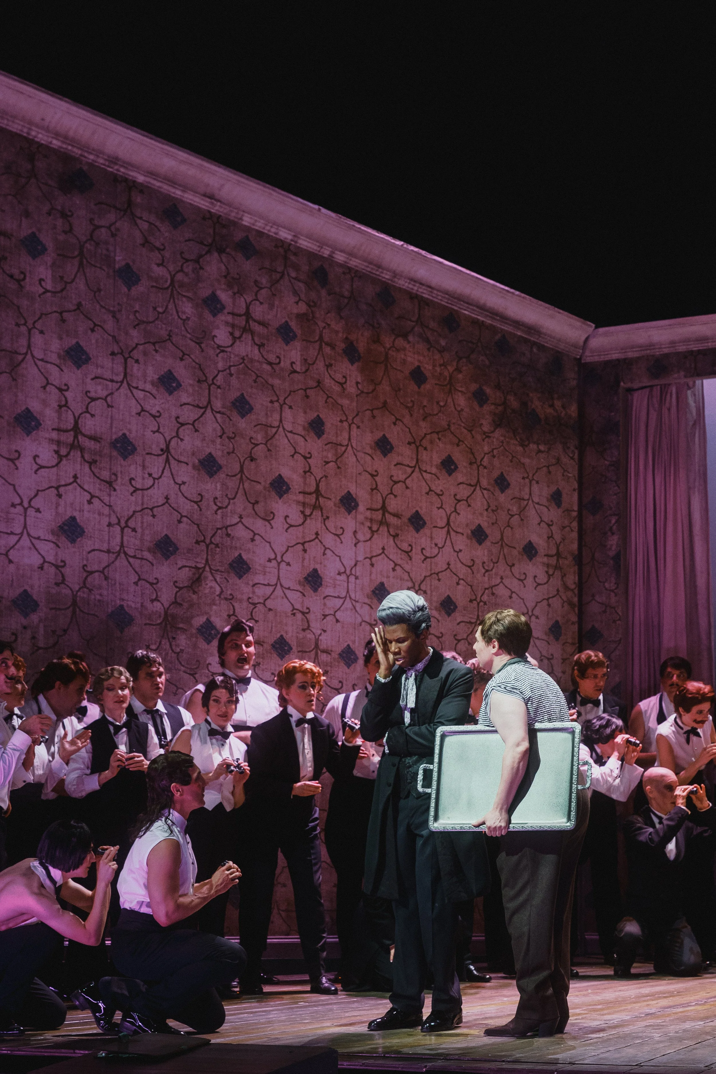 A theatrical stage scene with multiple performers. Two women in the foreground, one holding a large white case, the other with a hand on her face. In the background, a group of singers or performers dressed in formal costumes and dark vests appear to