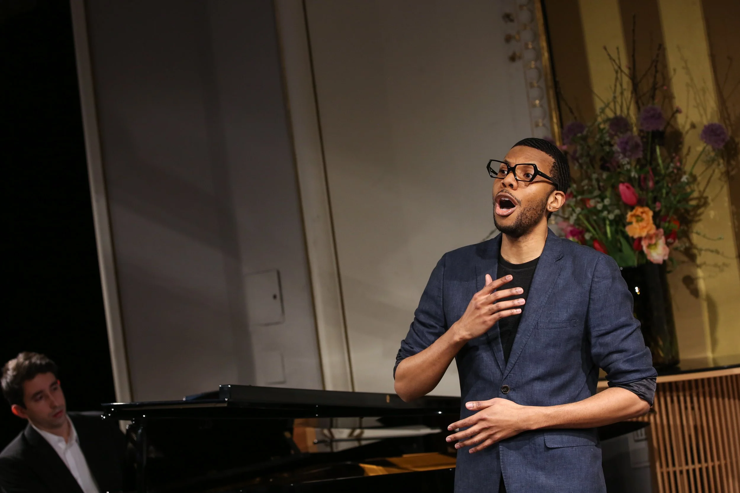 A man with glasses and a beard singing passionately with his hand on his chest during a performance, with a pianist playing in the background and a floral arrangement behind him.