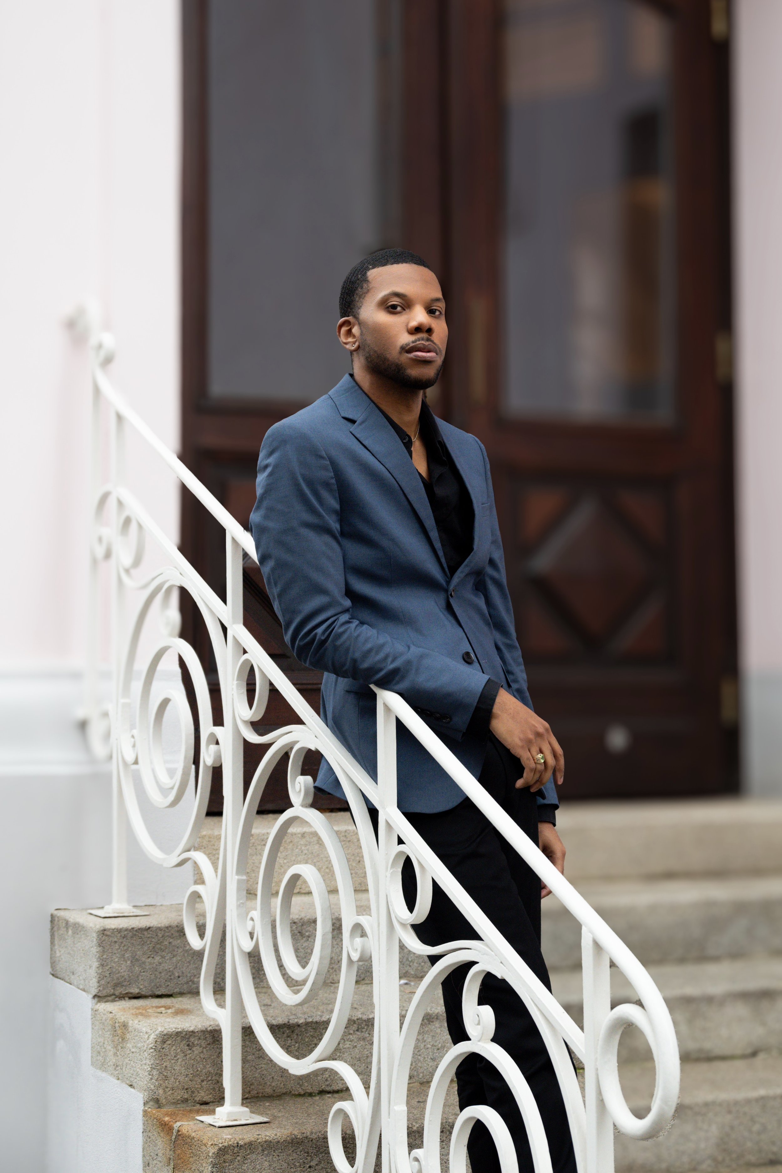 Aaron-Casey in a dark blue suit and black shirt standing on stairs outside a building with dark wooden doors, leaning on a white decorative railing.