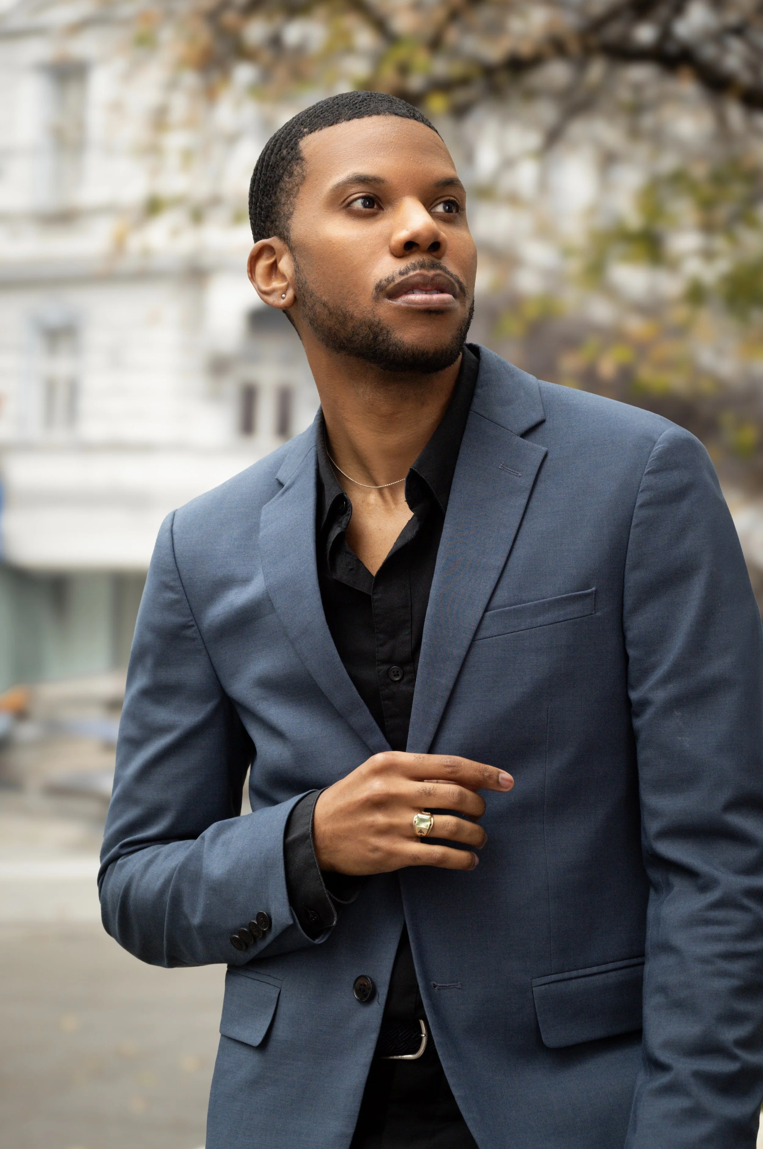 Aaron-Casey standing outdoors with blurred buildings and trees in the background. He has short hair, a beard, and is pointing with his right hand. He is wearing a gold ring on his finger and a small earring.