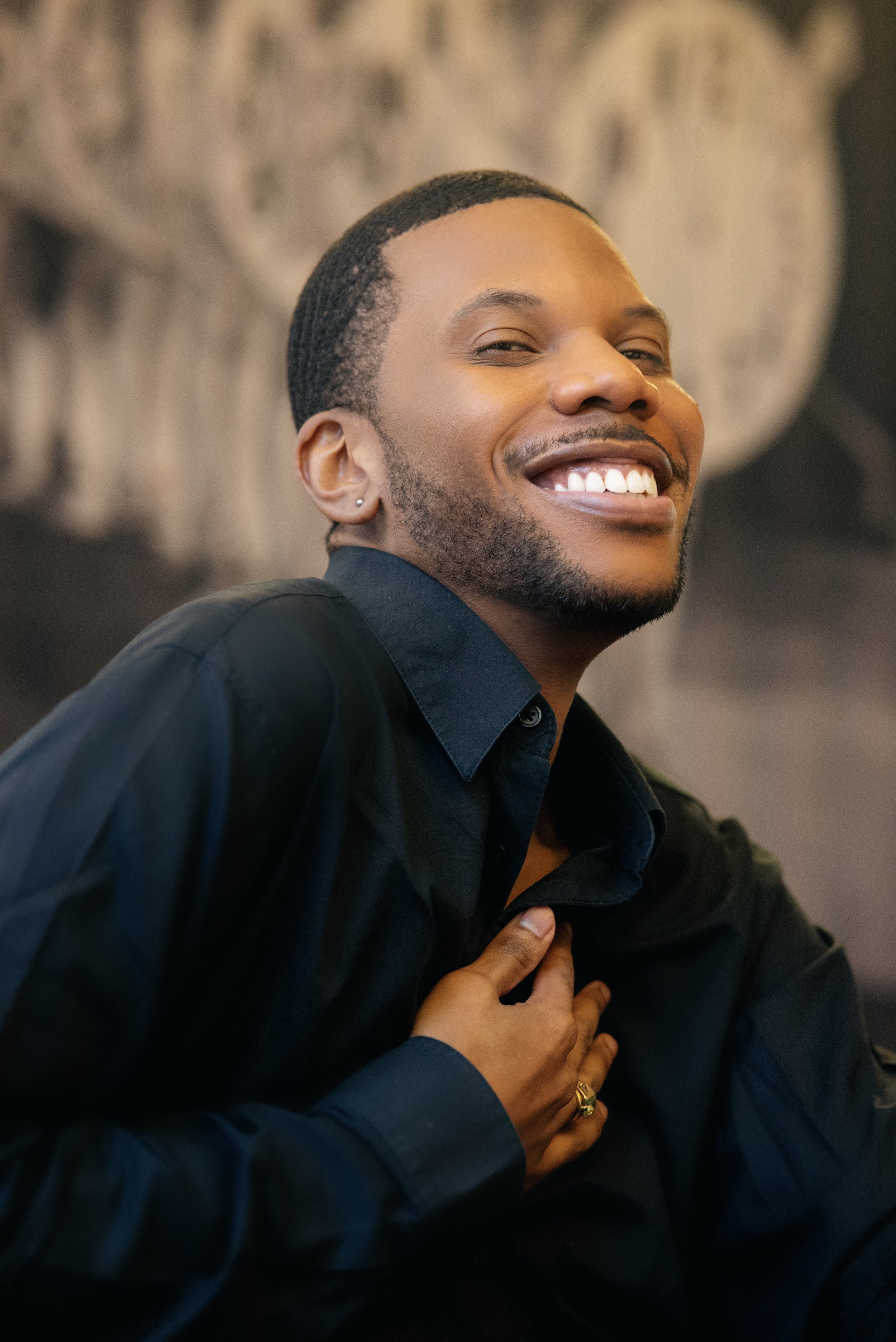 Aaron-Casey wearing a black shirt, with one hand on his chest, against a blurred background.