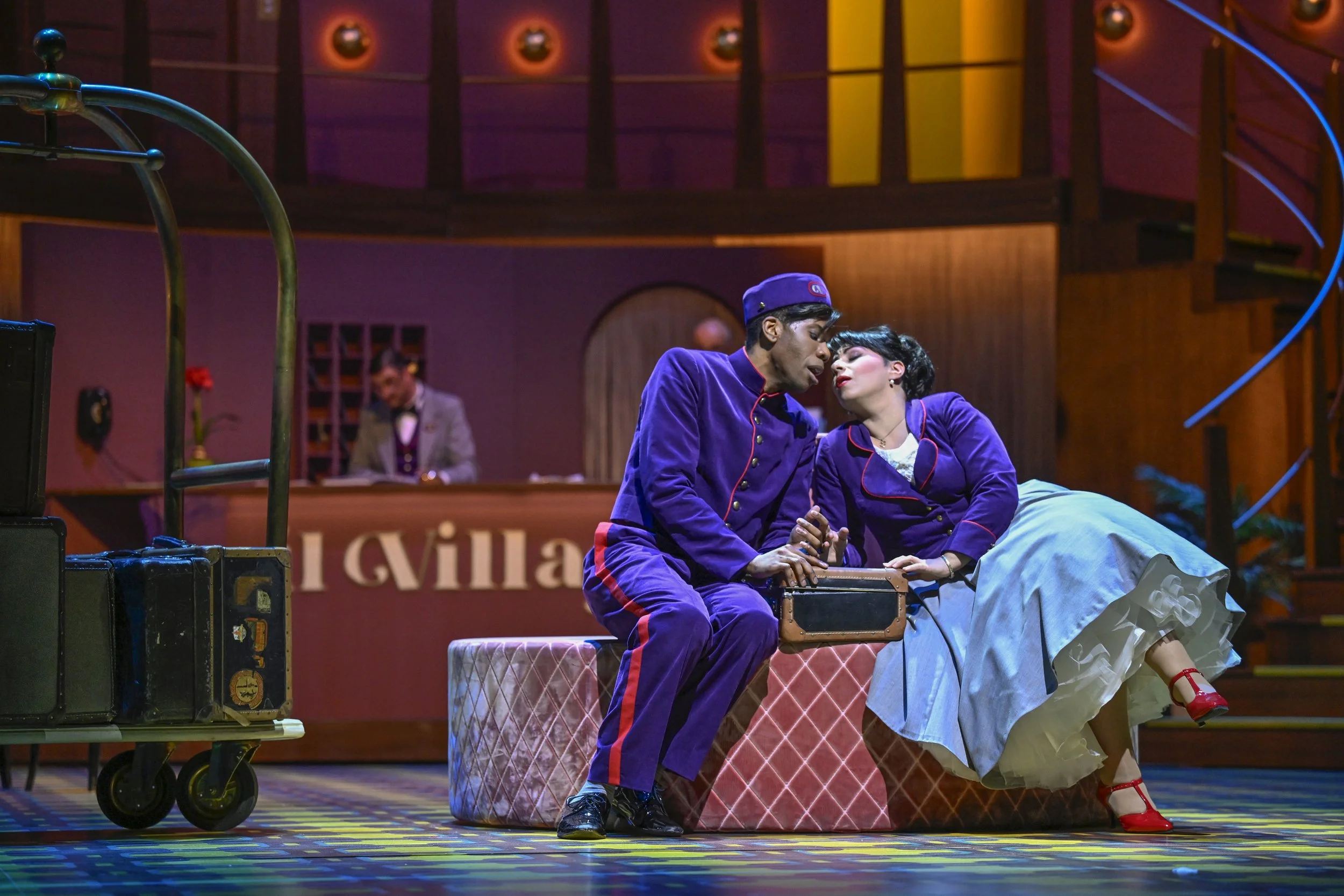 Two actors in vintage costumes performing a theatrical scene; a man in a purple uniform and a woman in a vintage dress sit closely on a pink checkered ottoman, leaning towards each other, while a man in the background looks on. The scene is set in a 