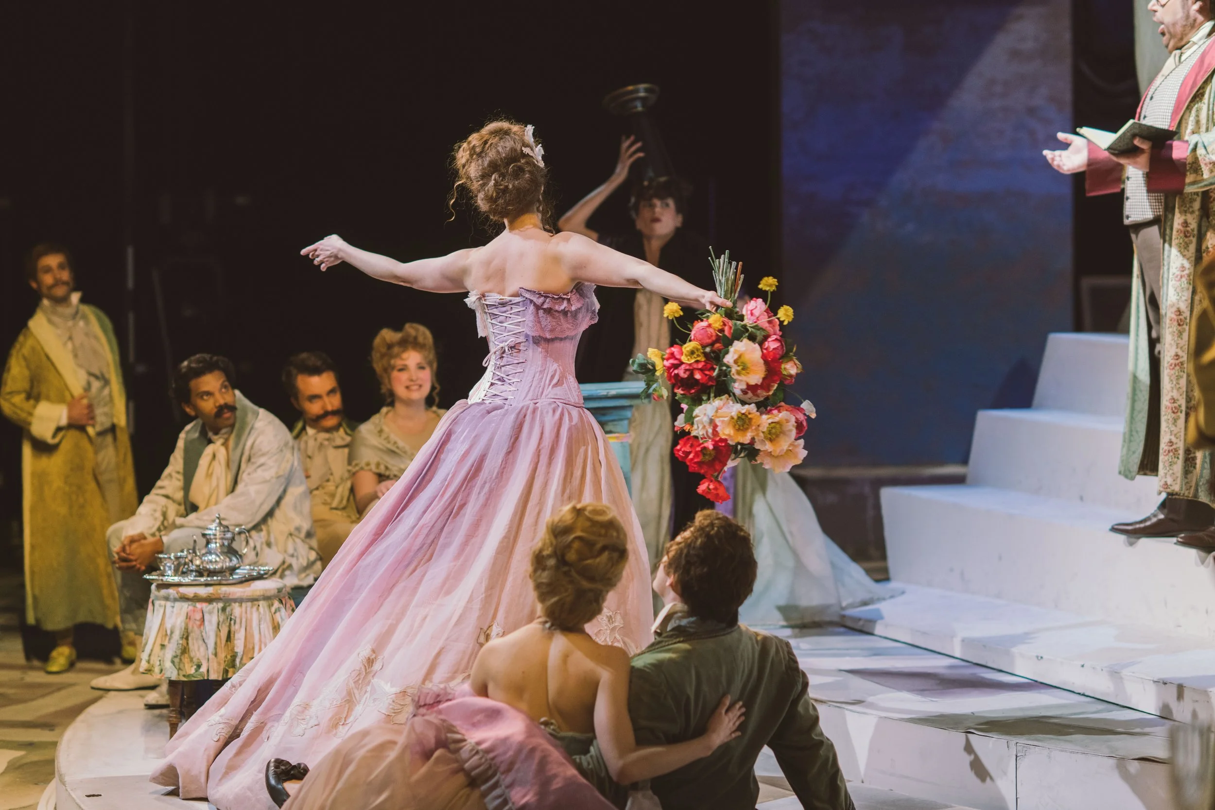 The image depicts a theatrical scene with a woman in a pink gown with a corset and lace-up back, holding a bouquet of flowers, standing on a stage with her arms outstretched. Several actors in period costumes are seated and standing around her, and t