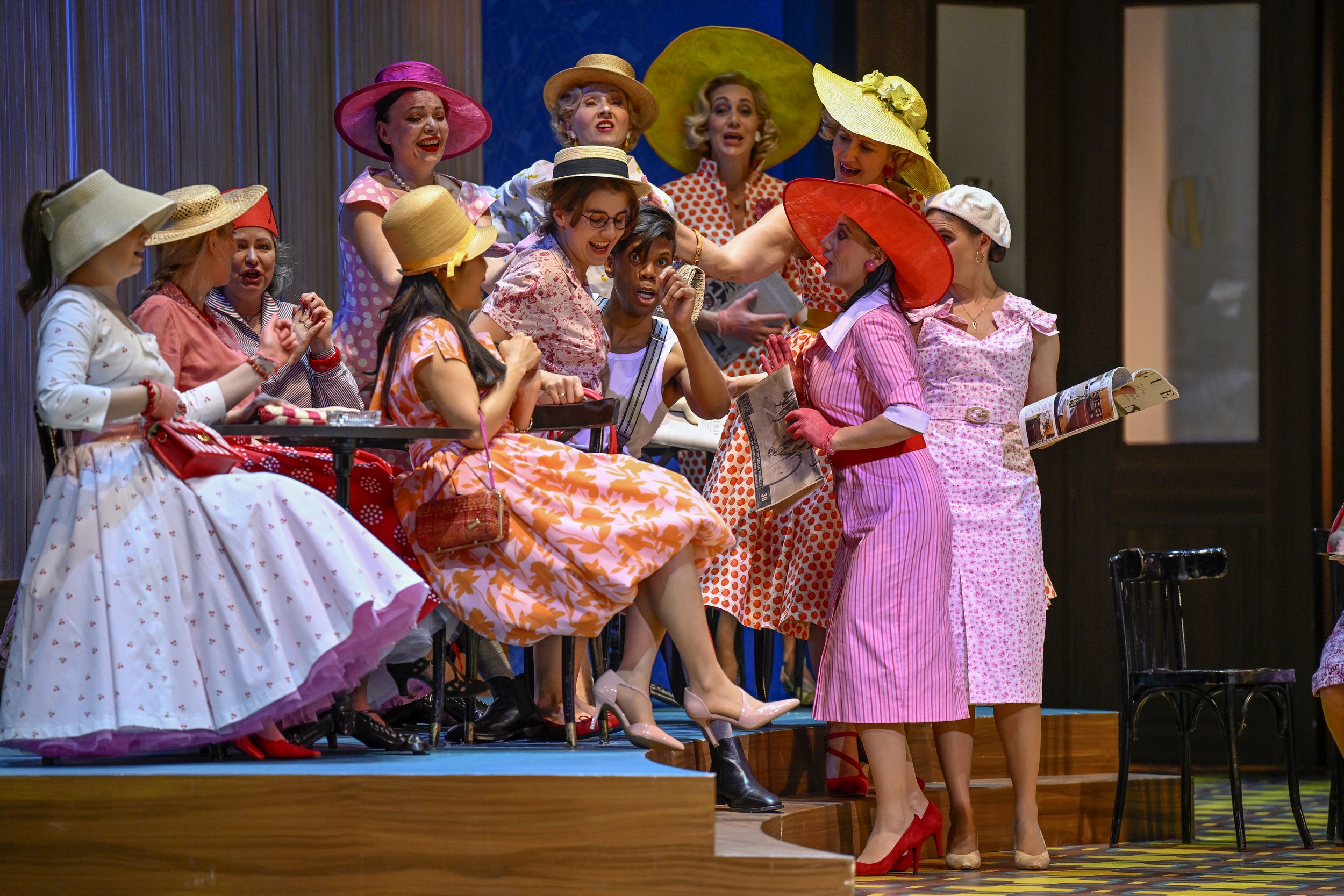 Group of women dressed in colorful vintage dresses and wide-brimmed hats, engaging in a lively conversation on a stage, some sitting and some standing.